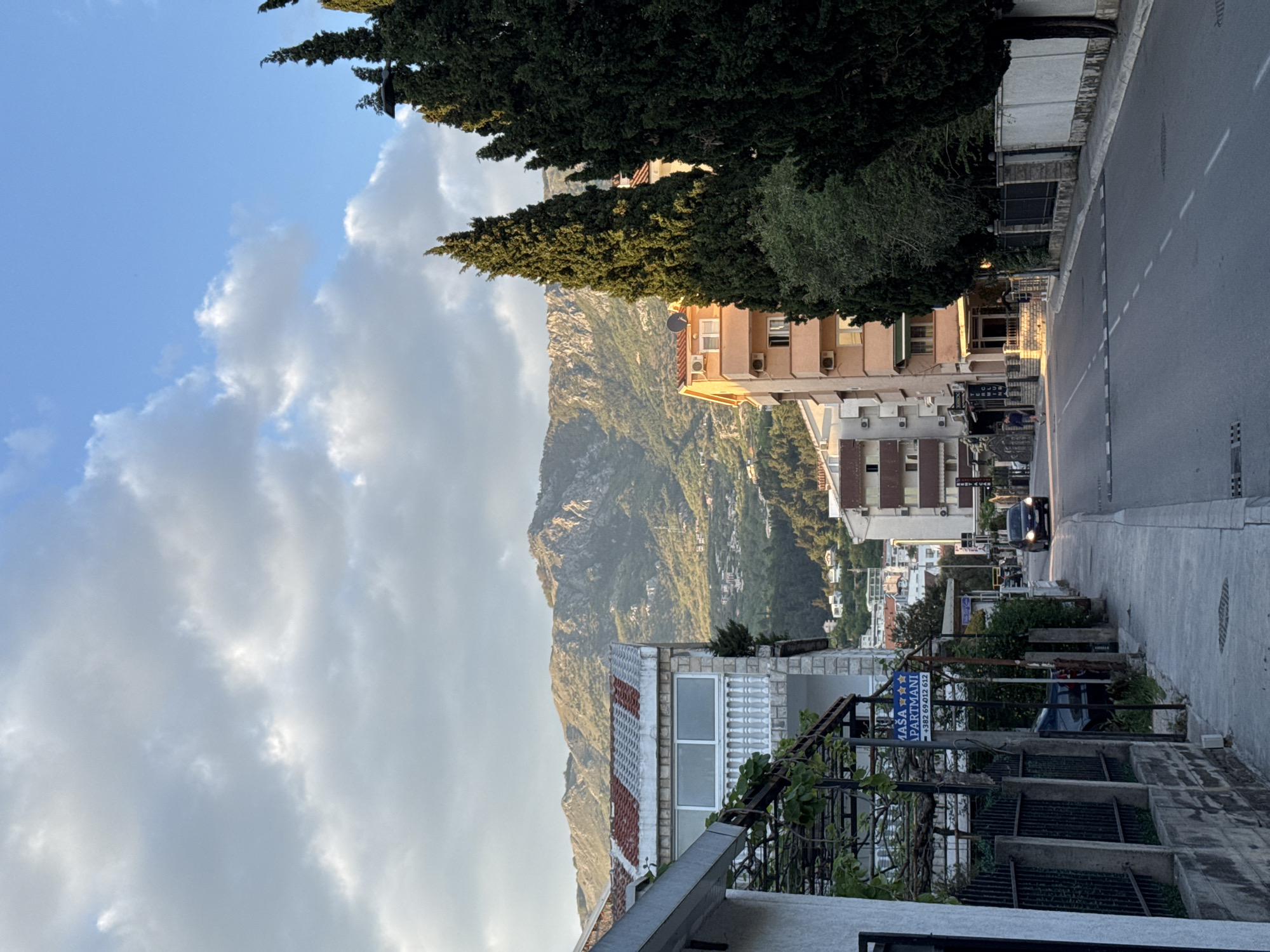 The image depicts a street scene in Budva, Montenegro. In the foreground, there is a sidewalk on the left side and a road on the right. Buildings line the street, including apartments and a business with a sign that reads "MAŠA Apartmani". A car is visible further down the road. In the background, a large mountain rises, partially obscured by trees and clouds. The sky is blue with white clouds, and the overall lighting suggests it's daytime. The camera angle captures the street receding into the distance.