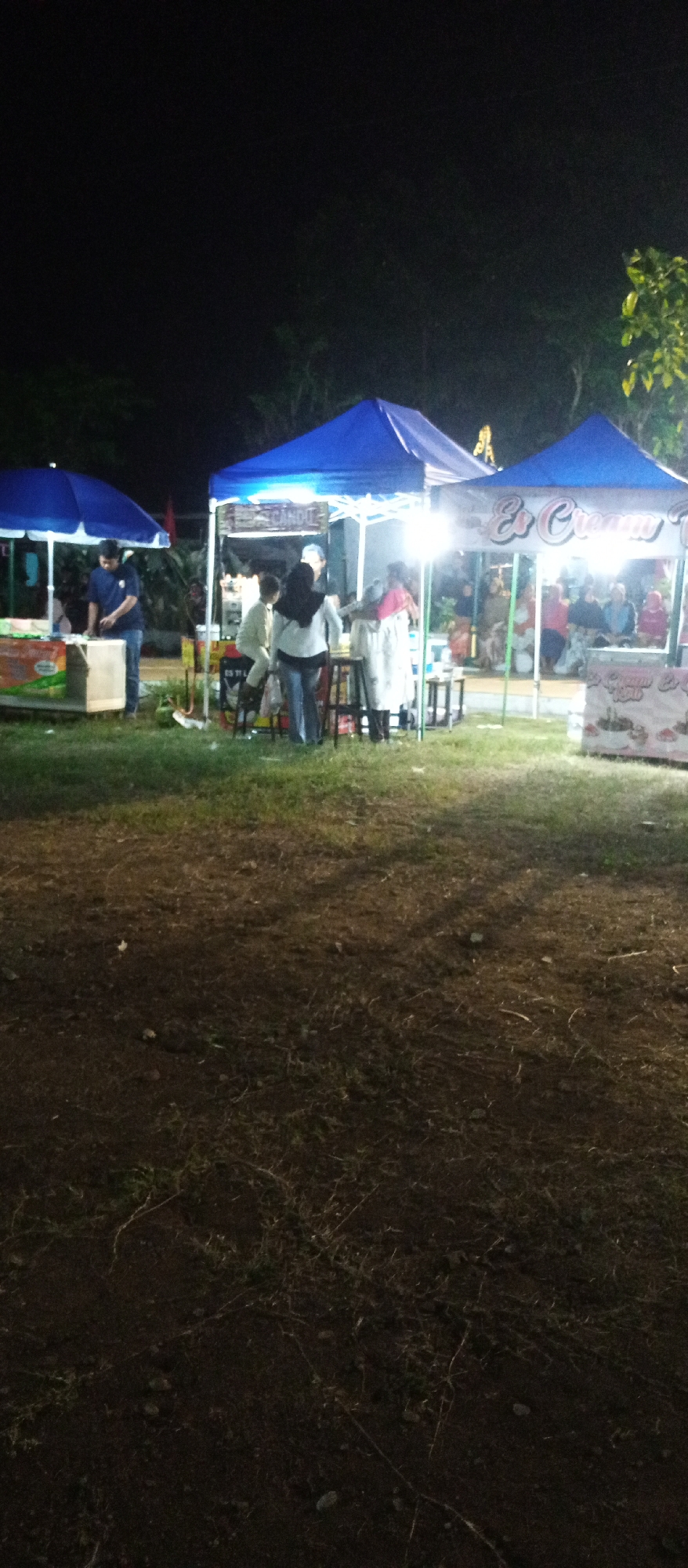 At night, several blue tents are set up on a grassy area with dark soil. In the foreground, the ground is covered with dry grass and dirt. In the middle ground, people are gathered around the tents, which appear to be food stalls. One tent has a sign that reads "Es Cream." A man in a blue shirt is standing at a table in front of a tent on the left. Several other people are standing or sitting at tables under the tents. In the background, trees are visible against the dark night sky. The scene suggests a bustling outdoor market or festival at night in Kaliwungu, Indonesia.
