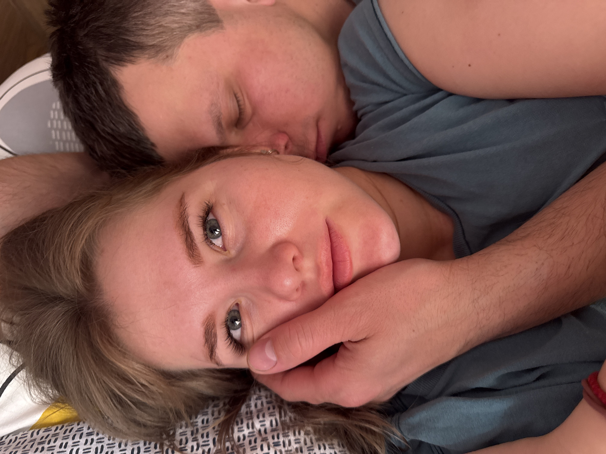 A man and a woman are lying in bed. The man is asleep with his eyes closed. The woman is awake, looking at the camera. A hand is touching the woman's face. The location is not evident from the image.