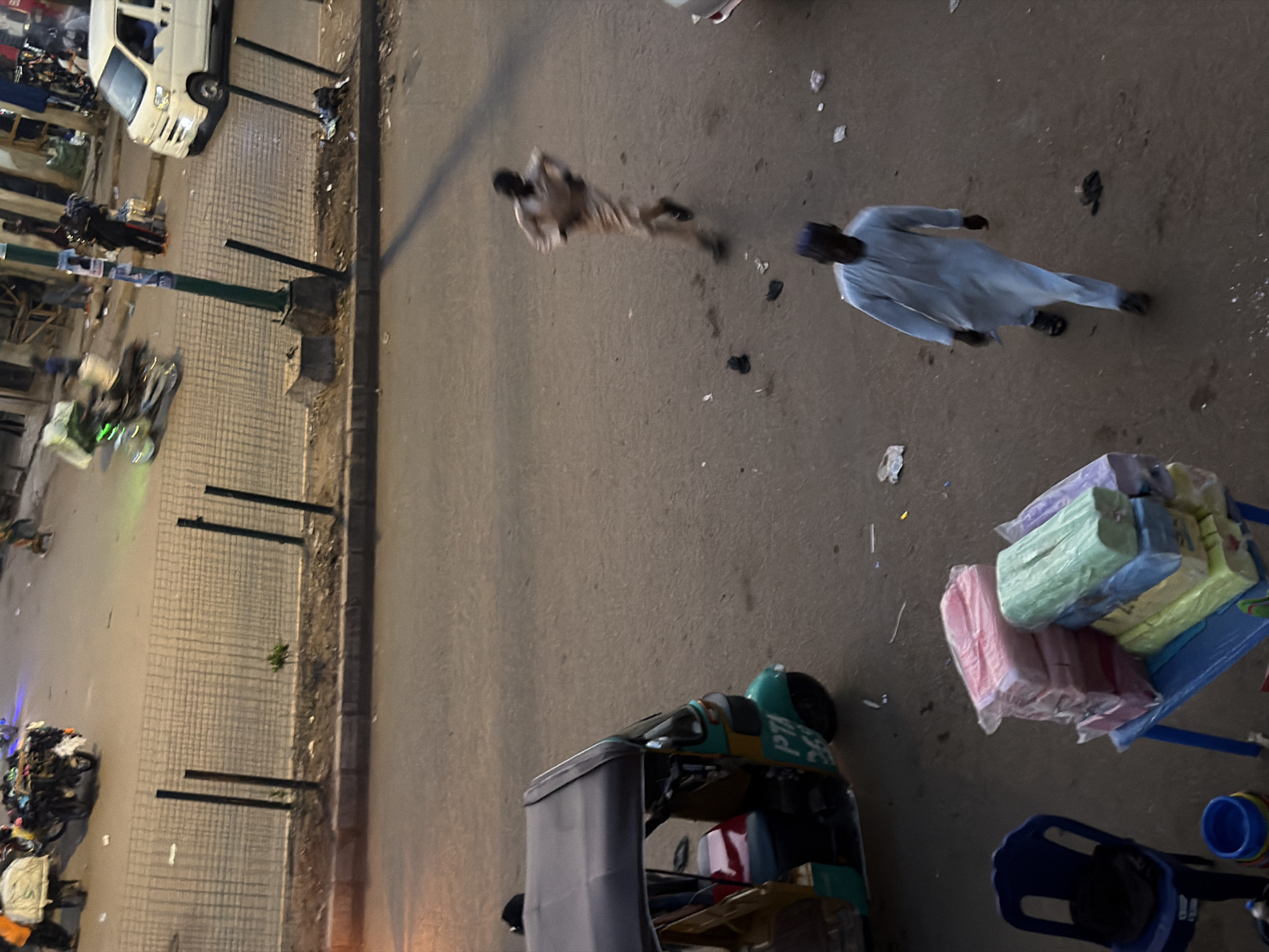 The image shows a street scene in Bukuru, Nigeria. In the foreground, a blue plastic chair is visible to the left. Next to it, a blue bucket and some stacked packaged goods in pink, green, blue, and yellow are displayed on a blue table. To the right and slightly behind, a man in a light blue traditional robe walks away from the camera. In the middle ground, an auto-rickshaw with a dark canopy is parked. Further back, a person in a light brown robe is running across the street. Beyond them, a white van is parked on the right, and a motorcycle with a cart loaded with goods is moving from left to right. The street is paved with cobblestones in some areas and asphalt in others. A metal fence runs along the side of the road, separating it from a raised dirt area with some sparse vegetation. The lighting suggests it is either early morning or late afternoon.