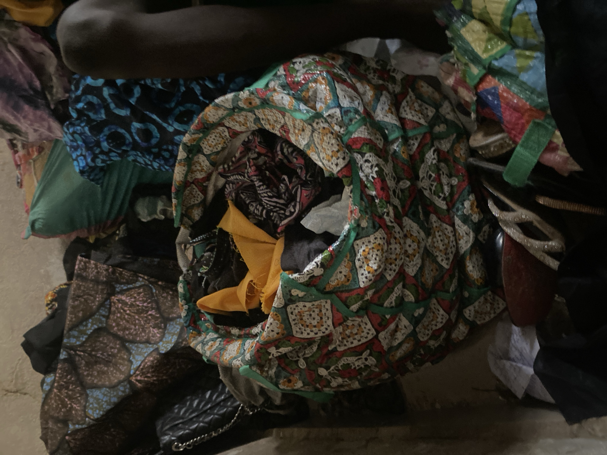 A medium shot captures a colorful, round fabric bag overflowing with clothing and other items in Jauro Yinu, Nigeria. The bag is patterned with white squares containing floral designs and red and yellow accents, trimmed with green fabric. Inside, a bright yellow piece of fabric is prominently visible, along with other garments in dark and patterned colors. To the left of the bag, a black quilted purse with a chain strap rests against a pile of fabrics. In the background, more clothing and textiles are visible, including a teal fabric and a blue and black patterned cloth. To the right, a dark-skinned person's knee and lower leg are visible, suggesting they are sitting or kneeling. The environment appears to be indoors, with a rough, textured wall in the upper left corner. Several other items, including what appears to be a shoe and folded colorful fabric, are scattered around the base of the bag.