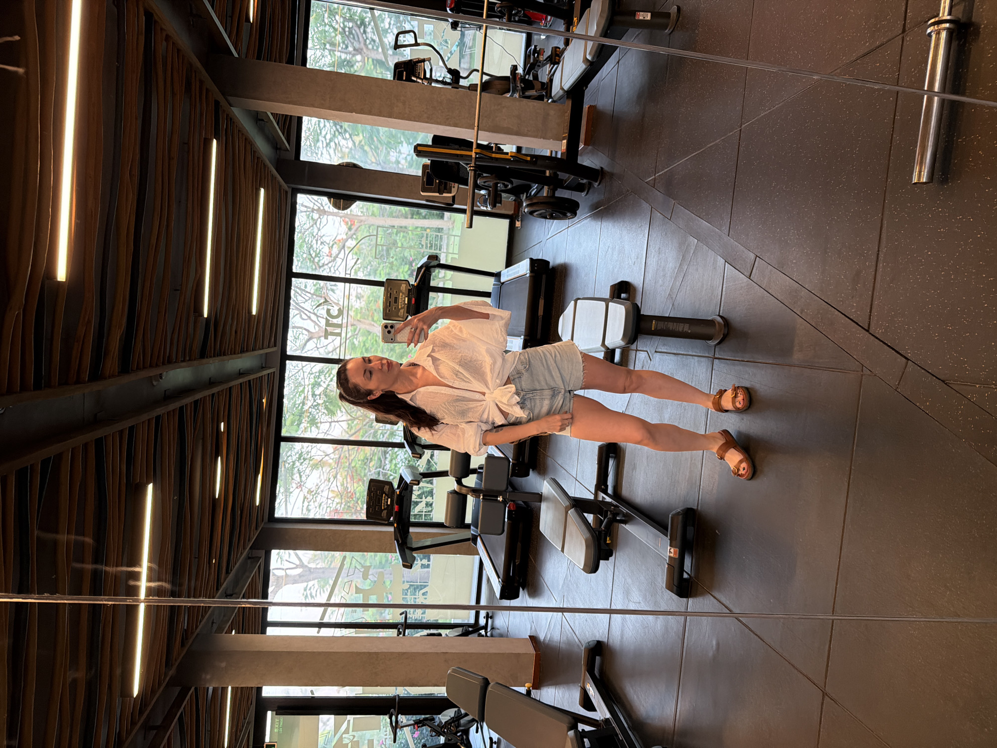 The image depicts a woman taking a mirror selfie inside a fitness center located in Dong Ninh Hoa Ward, Vietnam. The location is indicated by the "TTC" logo visible on the exterior through the gym's windows, and the tropical vegetation outside.
The woman, with dark hair tied back, stands in the center foreground, facing her reflection. She wears a white, collared, long-sleeved shirt tied at the waist, light blue denim shorts with frayed hems, and brown leather sandals. Her toenails are painted red. She holds a silver smartphone in her right hand, capturing her reflection. A ring is visible on her left ring finger.
The gym features dark grey interlocking rubber flooring. Structural columns are light grey, and the ceiling is characterized by dark, undulating wooden panels interspersed with linear fluorescent light fixtures and exposed dark metal beams. Large, full-height windows extend along the back and right sides, offering a view of abundant green trees, some bearing red flowers, typical of a tropical environment.
Reflected in the mirror, various gym equipment is visible, including at least two treadmills, several adjustable utility benches (one prominently in the mid-ground), a black abdominal crunch machine, weight racks with barbells, and an exercise bike partially seen on the left. A barbell lies on the floor in the lower right area of the reflection. The vertical edge of the mirror runs down the left side of the frame.