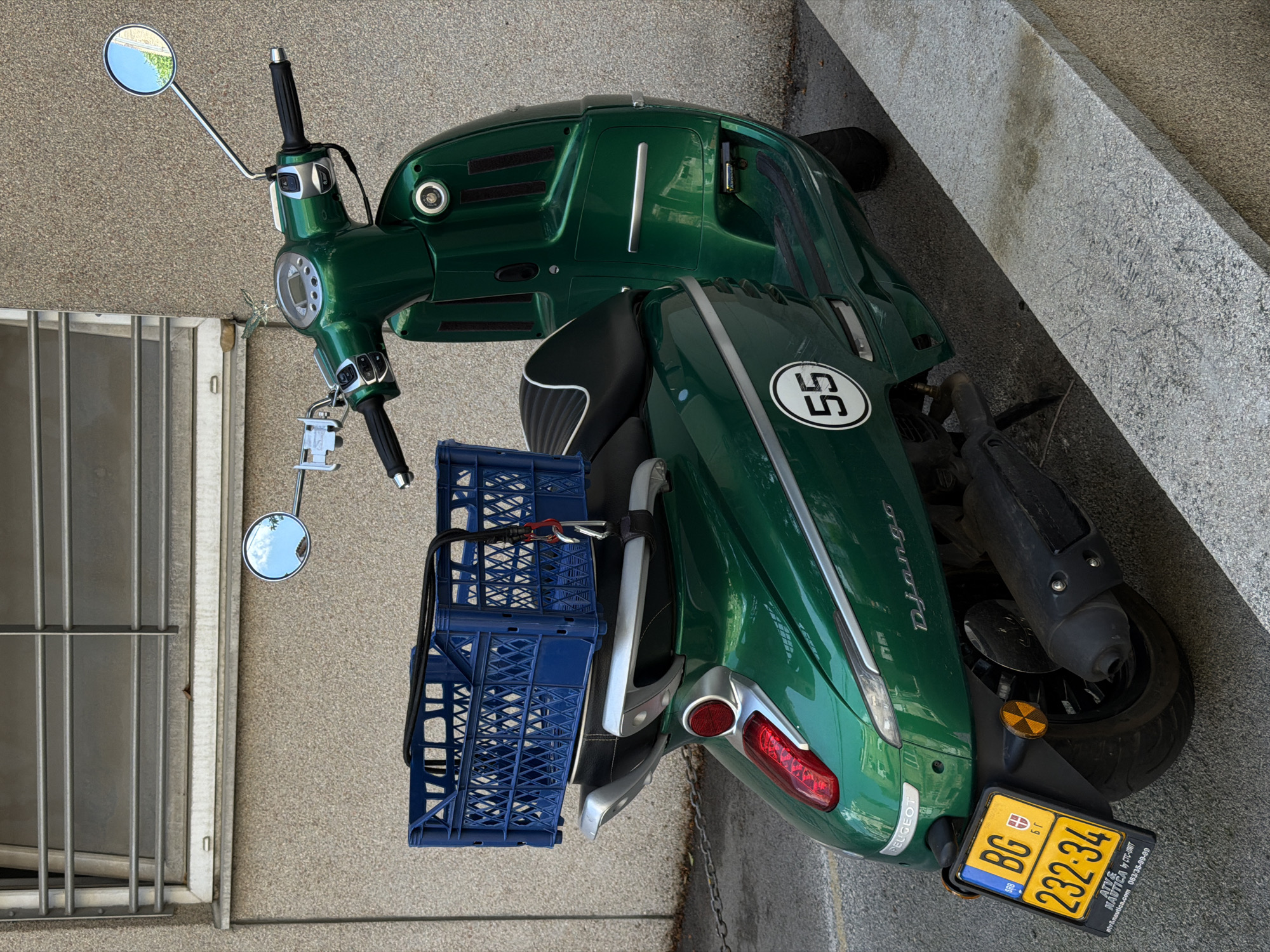 A green Peugeot Django scooter with a license plate from Belgrade, Serbia, is parked on a concrete surface next to a building with metal bars covering a window. The license plate reads "BG 232-34". A blue plastic crate is attached to the back of the scooter. The scooter has the number "55" displayed on its side. The scooter is angled diagonally in the frame.