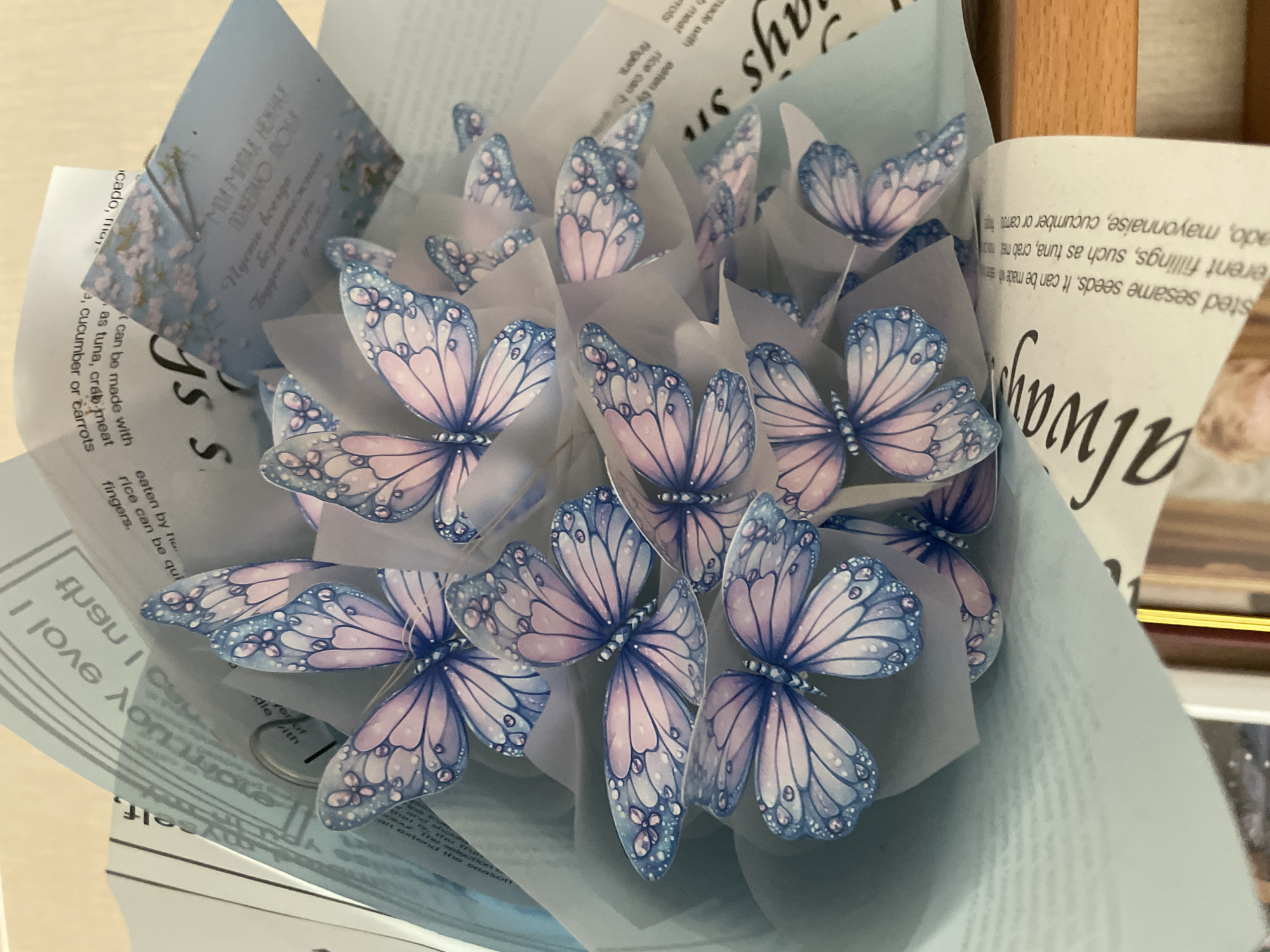 This is a close-up, eye-level, indoor shot of a bouquet decorated with numerous artificial butterflies. The butterflies are predominantly blue and pink with delicate patterns and what appear to be water droplets. They are attached to thin wires and are arranged in a cluster, some overlapping others. The bouquet is wrapped in light blue and white paper, with text visible on some of the layers. The text appears to be in English and some of it is upside down. One piece of paper, possibly a card, has blue floral imagery and writing in a light blue font. The background is blurred but suggests a wooden surface, possibly a table, and another blurred object that might be a bookshelf. The lighting is soft and natural. The image does not provide any information about the location within a city or any specific environmental details beyond being indoors.