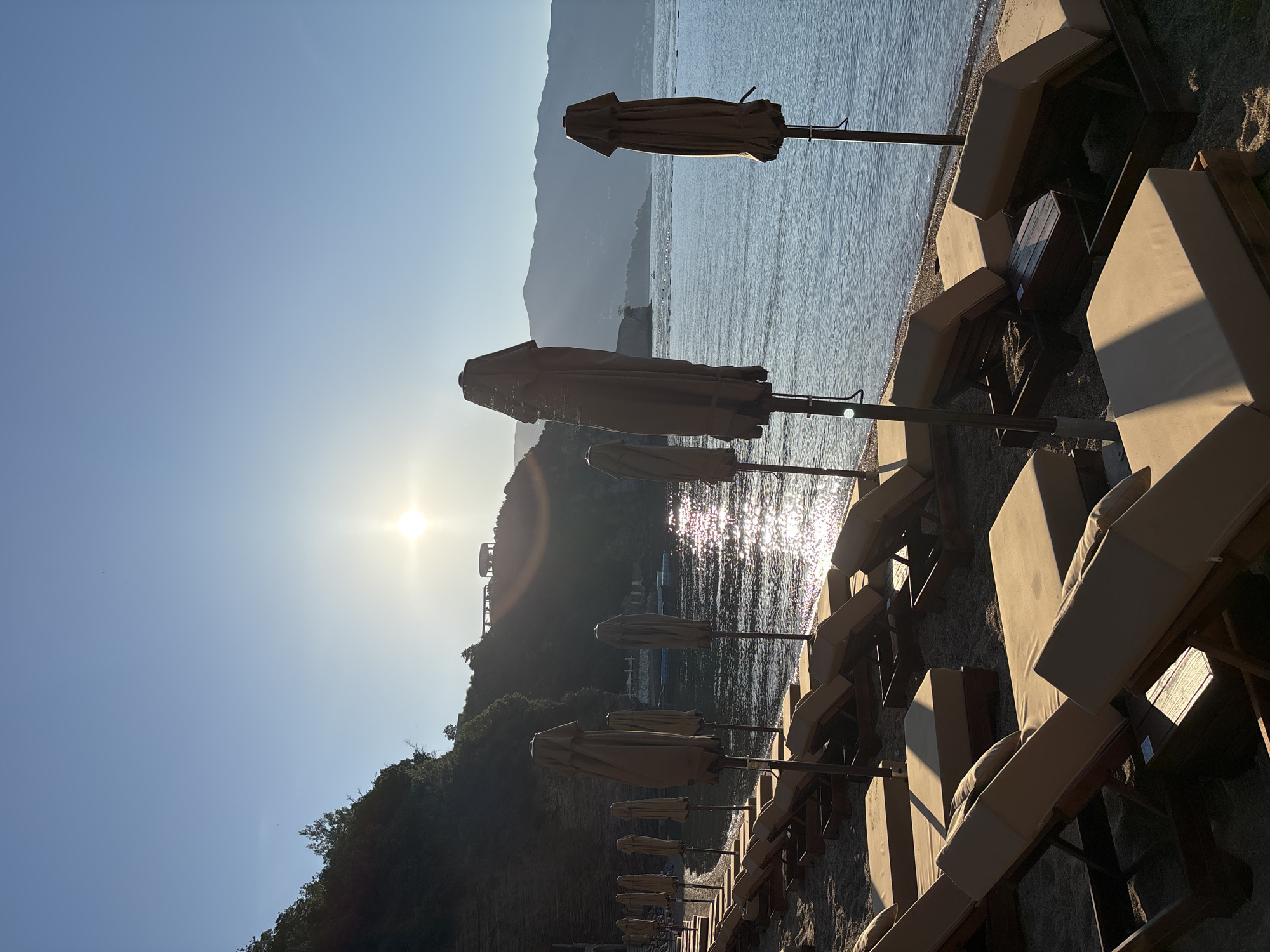 The image shows a serene beach scene at dawn or dusk. The sun is low in the sky, casting a warm, golden light across the water and the shore. A line of empty lounge chairs and closed umbrellas stretches along the sandy beach, suggesting a quiet moment before or after a busy day. The calm sea reflects the sunlight, creating a shimmering effect. In the background, a lush green hillside rises from the coastline, topped with a few structures. The overall atmosphere is peaceful and tranquil.
