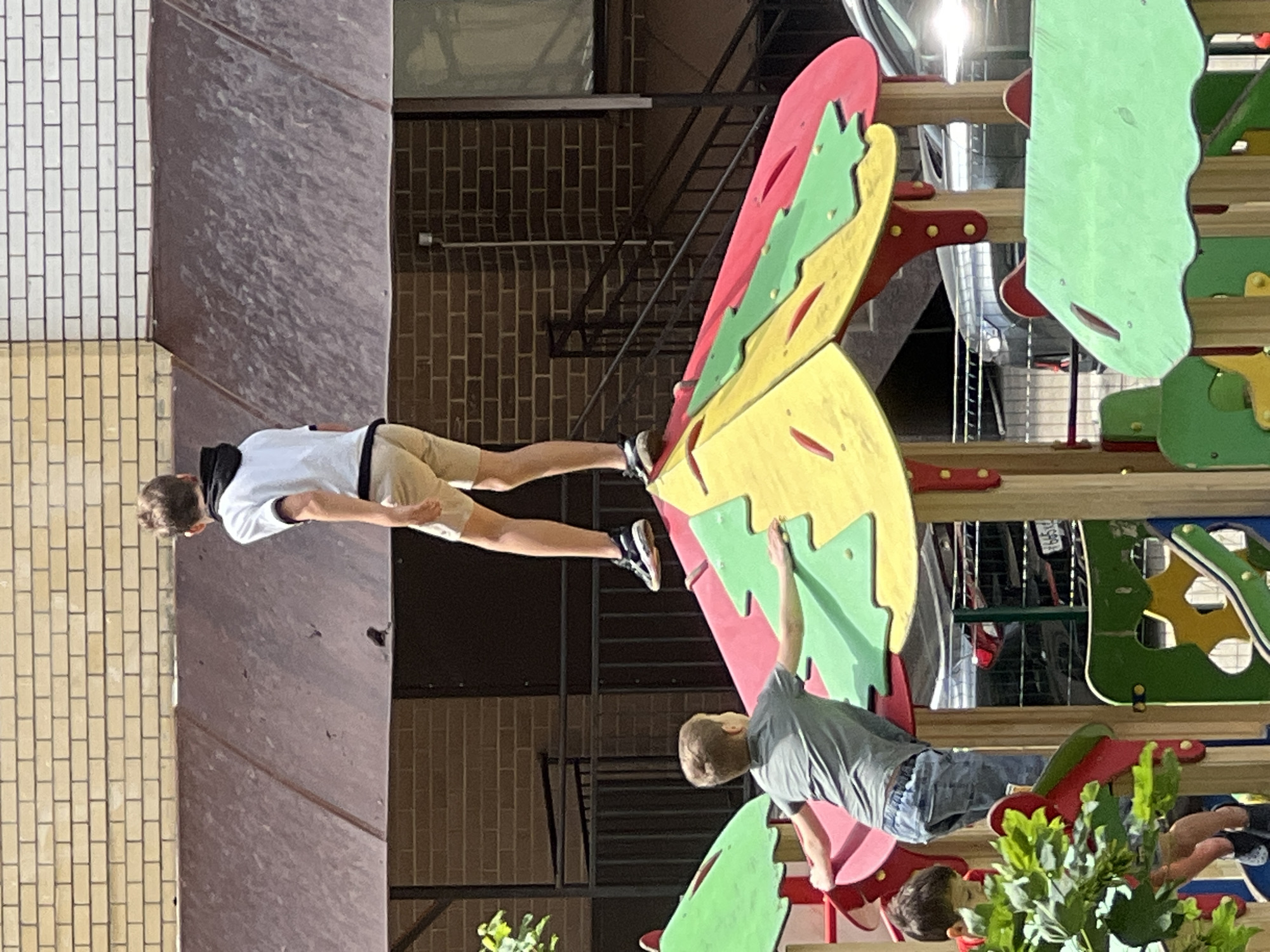 Two boys are on a colorful butterfly-shaped playground structure in an outdoor setting. One boy is wearing a white t-shirt, khaki shorts, and sneakers. He is reaching towards the top of the butterfly. The other boy is wearing a gray t-shirt and blue jeans and is positioned near the bottom. A third boy is partially visible near the base of the structure, obscured by foliage. The background shows a brick building, a metal staircase, and other parts of the playground. The location is Krasnodar, Russia.