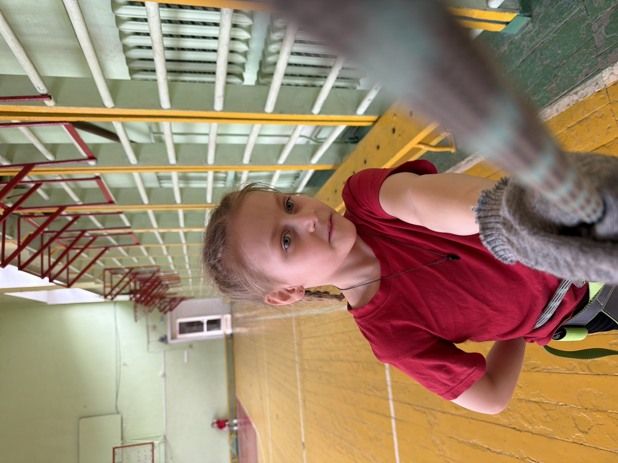 This image, taken in Izhevsk, Russia, captures a young girl engaged in an indoor rope activity within what appears to be a gymnasium or sports hall. The girl, likely of primary school age, has light hair styled in braids and light eyes. She is wearing a red short-sleeved t-shirt and a multi-colored climbing harness with prominent green and black straps. Her right hand, possibly gloved or wrapped, firmly grasps a thick rope that extends out of view. She is looking directly at the camera with a calm, neutral expression.
The setting features a worn, yellow-orange floor marked with painted lines, characteristic of sports courts. The walls are painted a light green. Above, the ceiling structure reveals exposed beams and a series of white horizontal bars, suggesting gym wall bars or part of a larger climbing system. In the background, red metal climbing frames or safety structures are visible along the walls. A window and what appears to be a fire extinguisher are also discernible on a distant wall. Faintly visible in the deeper background is a basketball hoop or backboard. The overall lighting suggests it is daytime. No legible text is visible in the image.