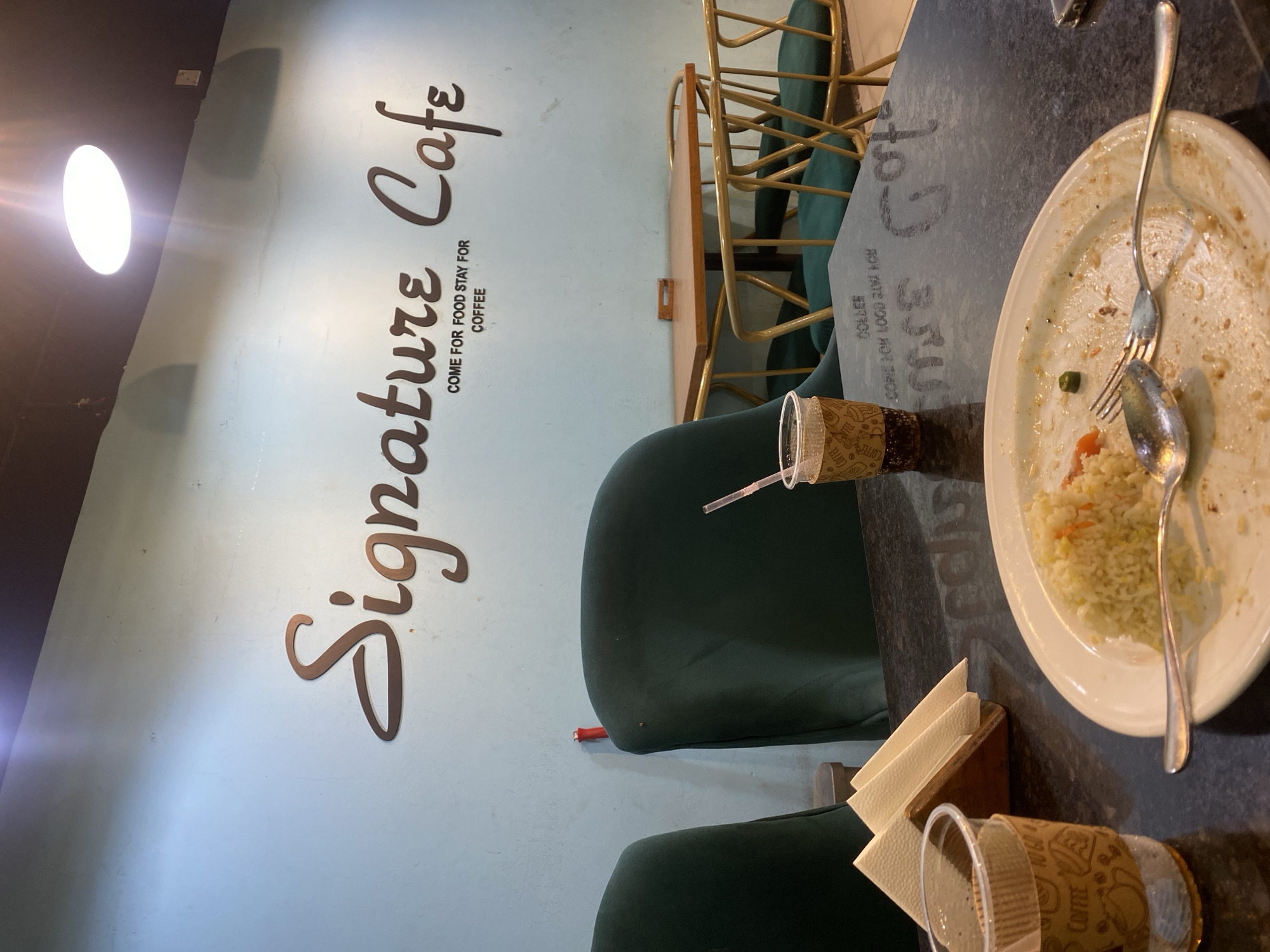 The image depicts the interior of a cafe, likely located in Dhaka, Bangladesh. On the light blue wall, a stylized sign reads "Signature Cafe" with the tagline "COME FOR FOOD STAY FOR COFFEE." In the foreground, a dark, speckled table holds the remnants of a meal. A white plate contains a small amount of fried rice, with a fork and spoon resting on it. To the left of the plate, a disposable coffee cup with a straw is partially visible, and a wooden napkin holder with white napkins is situated behind it. In the background, two dark green upholstered chairs are visible, one of which has a red object on its back. Beyond these chairs, a small wooden table with gold-colored chairs is set up. The overall ambiance suggests a casual dining or coffee shop setting.