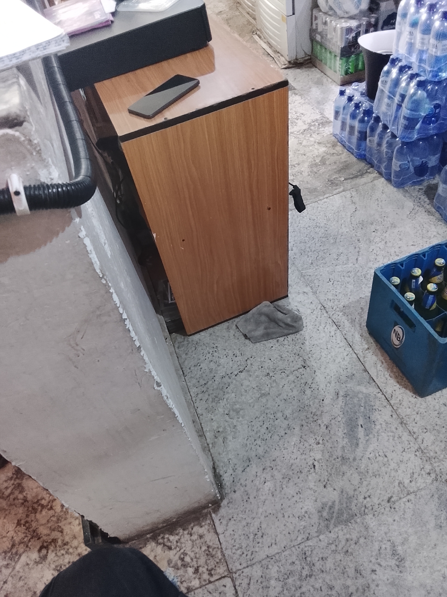 The image shows an indoor scene, possibly a storage area or a shop, with a focus on a wooden counter and the floor. On the counter, a black smartphone is placed horizontally, with its screen facing upwards. To the left of the counter, a black ribbed hose is attached to a white fixture.
In the background, to the right of the counter, several cases of bottled water are stacked, predominantly in clear plastic wrapping. Adjacent to the water bottles, a blue plastic crate is filled with bottled beverages, likely beer, with visible labels. The floor is a speckled grey, consistent with commercial flooring. A grey cloth is visible on the floor near the base of the counter.
The scene appears to be set during daylight hours, suggested by the ambient lighting. There are no obvious indications of specific weather conditions. The overall impression is one of a functional, possibly commercial, space. There is no visible text that can be clearly read. The composition and focus suggest an observation of everyday objects within this setting.