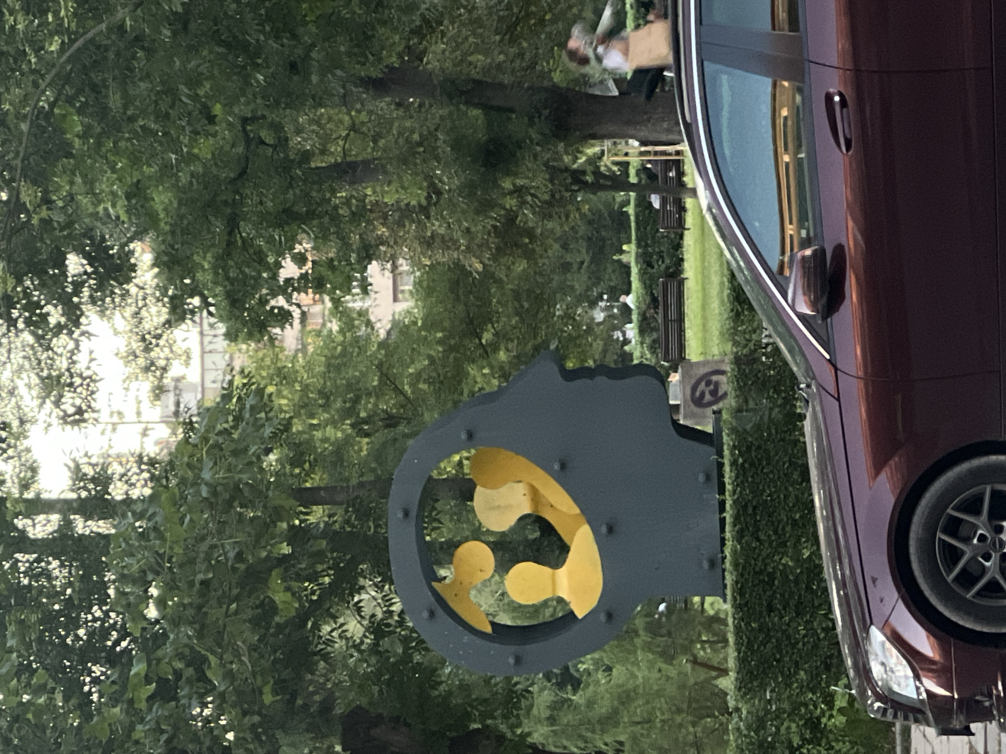 In Sofia, Bulgaria, a large, dark grey sculpture resembling a human head profile is displayed in a park setting. The sculpture features a circular opening within the head, containing several yellow figures that appear to represent people or abstract shapes. The background is filled with lush green trees, some of which have sunlight filtering through their leaves, suggesting it is daytime. In the middle ground, several park benches are visible, with a few individuals seated. A dark red car is prominently positioned in the foreground, partially obscuring the lower portion of the sculpture and the surrounding greenery. The car's front passenger side, including the headlight and wheel, is clearly visible. The scene depicts an outdoor public space with a blend of art, nature, and urban elements.
