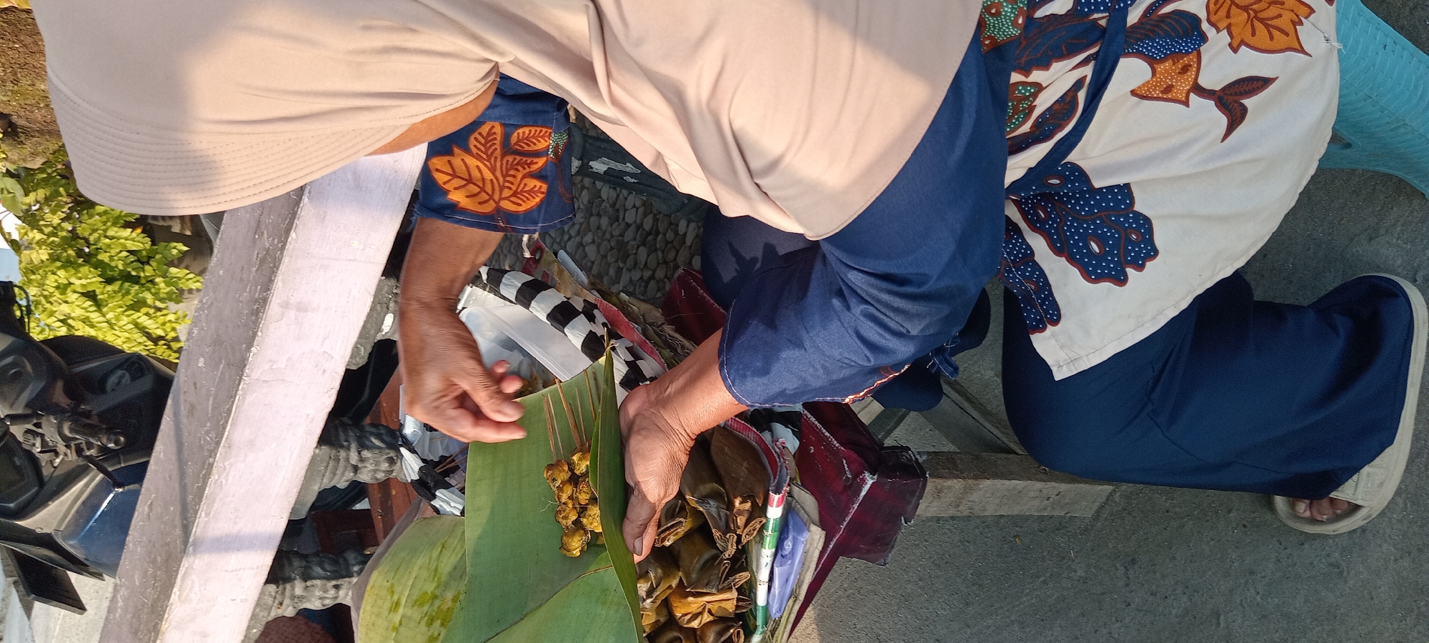 In Kalipucang, Indonesia, an individual wearing a beige hijab, a blue long-sleeved shirt with an orange leaf pattern on the sleeves, and dark blue pants is preparing food. They are seated on a light blue plastic chair, with their feet wearing light-colored sandals visible. The person's hands are in focus as they arrange skewers of what appear to be small pieces of food, possibly meat or seafood, on a large green banana leaf. In front of them, on a wooden table, are several bundles of food wrapped in banana leaves. The table is supported by a rough wooden beam. Behind the table, a motorcycle is partially visible, along with a potted plant with green foliage. The ground appears to be a mixture of concrete and gravel. The scene is illuminated by natural light, suggesting it is daytime.