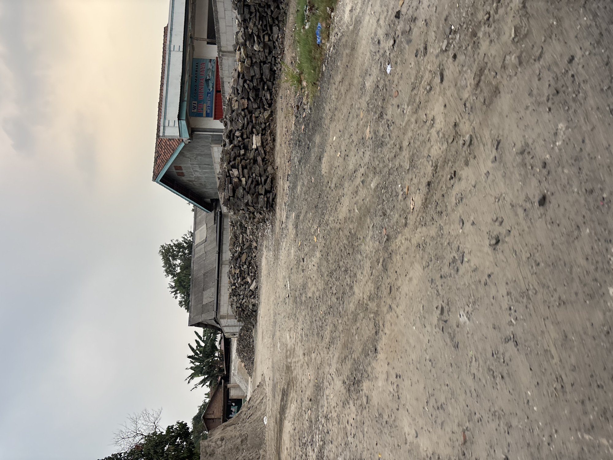 The image depicts an outdoor scene in what appears to be a semi-developed area in the city of Bekasi, Indonesia. In the foreground, a dirt road is visible, with piles of sand and stones on either side. In the midground, several buildings with corrugated metal roofs are present. One building has a sign that reads "WI TOKO MATRIAL PD. WINDA JAYA" with a phone number below. The sky is overcast with some hints of sunlight. The overall impression is of a construction or material supply yard.
