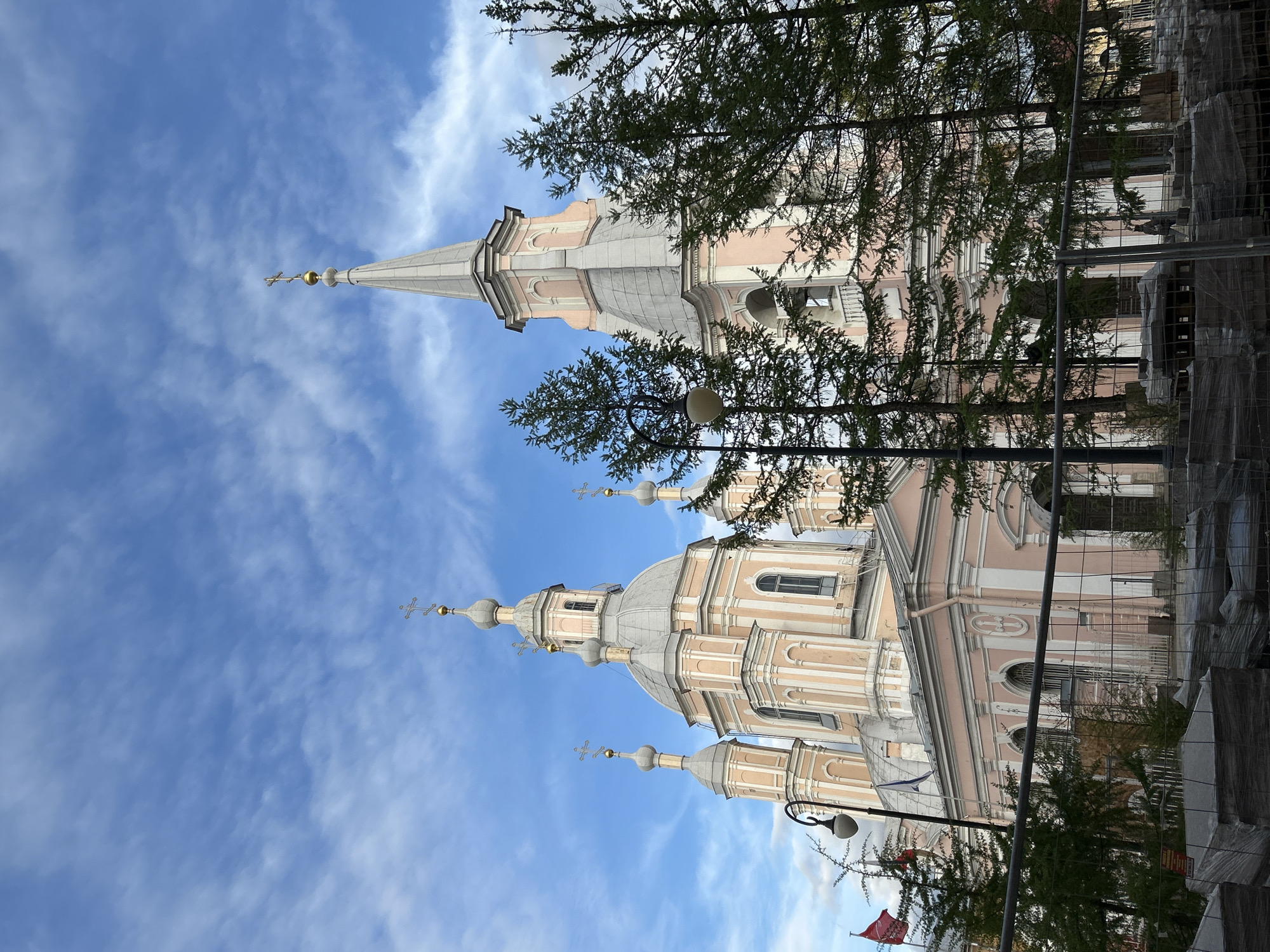 The image captures a low-angle perspective of the Smolny Cathedral in Saint Petersburg, Russia. The cathedral's ornate facade, domes, and spire rise against a backdrop of a bright blue sky with wispy clouds. The structure is characterized by its pale pink and white exterior, adorned with decorative columns, arched windows, and gilded crosses atop its domes. In the foreground, tree branches partially obscure the view, adding depth to the composition. Construction scaffolding can be seen on the right side of the image. A street lamp is visible at the bottom of the image on the right side.