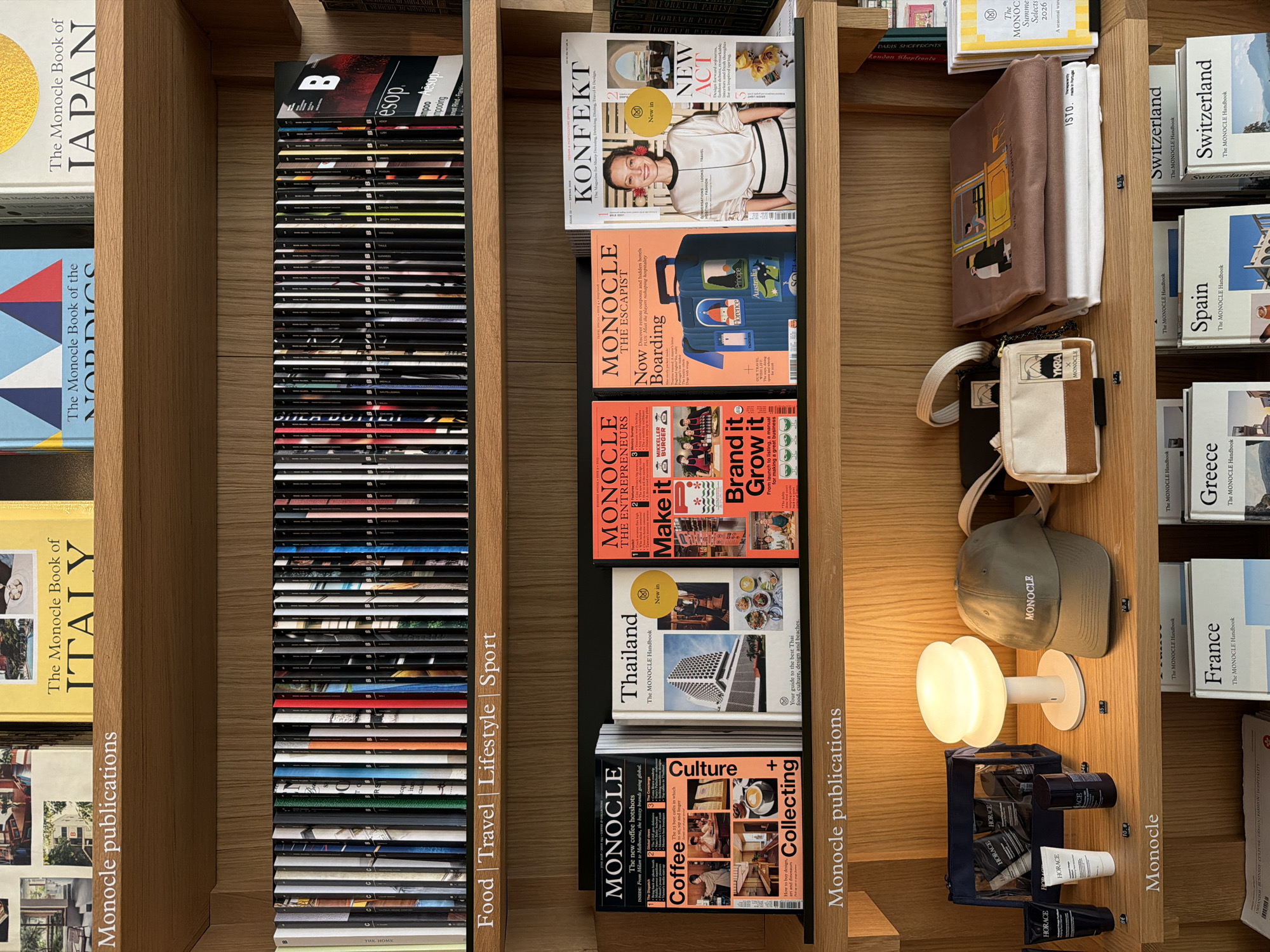 Here is a detailed description of the image:
**General Description:**
The image shows a wooden bookshelf filled with various Monocle publications and related items. The organization of the shelves suggests a curated display of products.
**Specific Details:**
* **Shelves and Publications:** The bookshelf has multiple shelves each dedicated to different categories of Monocle publications:
* Books on countries or regions, such as Japan, Italy, the Nordics, Switzerland, Spain, and Greece.
* Magazines related to food, travel, lifestyle, and sports.
* Magazines focused on entrepreneurship, culture, and design.
* Handbooks.
* **Magazines and Books:** Titles like "KONFEKT," "MONOCLE the ESCAPIST," "Thailand The MONOCLE Handbook," and "MONOCLE Coffee + Collecting" are visible. There are also numerous issues of Monocle magazine stacked vertically.
* **Other Items:**
* A small lamp providing light on the shelf.
* A Monocle-branded hat.
* A collection of Horace grooming products.
* A couple of bags or pouches with Monocle branding.
* **Branding and Labels:** The Monocle brand is prominently displayed on various items. There are also labels and price tags on some products indicating they may be for sale.
**Location:**
It looks like the items may be sold near the Monocle shop in Paris because there is a Monocle book on Paris.