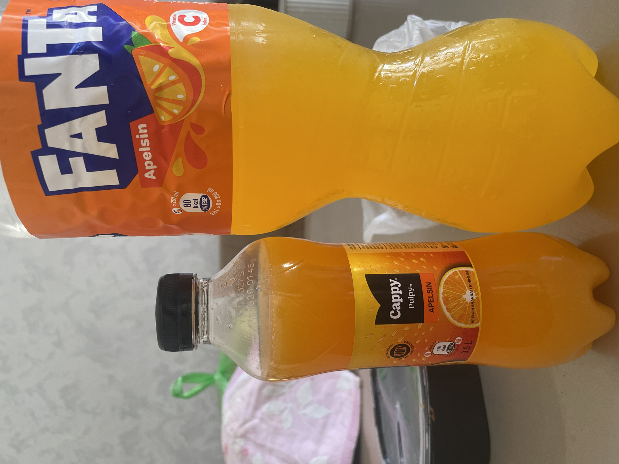 The image shows two orange-colored beverages, likely fruit juices or sodas, on a countertop. One is a smaller, clear plastic bottle with a black cap and an orange label. The label is partially visible, displaying the brand "Cappy" and the flavor "Pulpy™ APELSIN" (orange). The other is a larger, orange plastic bottle with a white label featuring the "FANTA" logo in blue and orange. The "FANTA" label also indicates "Apelsin" (orange) flavor and displays nutritional information. The background is blurred but suggests an indoor setting with a textured wall and some light-colored fabric or plastic. There is no indication of the location being in Tashkent, Uzbekistan.