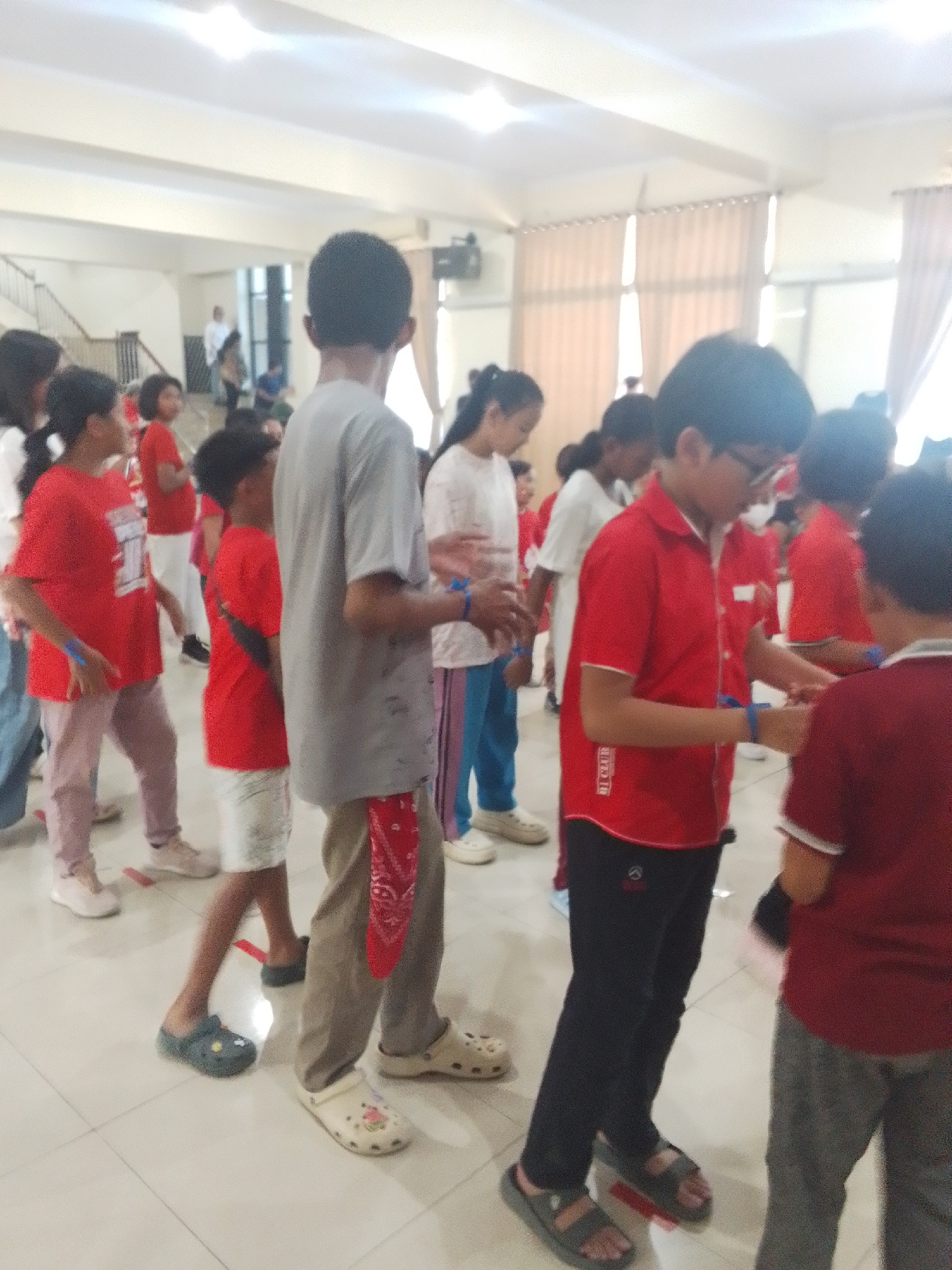 In a well-lit indoor space, possibly a community hall or school gymnasium in Makassar, Indonesia, a group of children and teenagers are gathered. The floor is tiled in a light color. Many are wearing red shirts, suggesting a uniform or team event. Some are engaged in an activity, indicated by their focused expressions and interactions. One boy in the foreground wears a red collared shirt and black pants, with glasses and a serious expression. To his right, another boy in a maroon shirt with white trim is partially visible. In the center of the image, a boy in a grey t-shirt with a red bandana tied around his waist stands facing away from the camera. Other individuals, mostly young, are dispersed throughout the space, some in red shirts and others in casual attire. The background shows curtains and fluorescent lighting, typical of an indoor event venue. The overall atmosphere appears to be one of organized activity or learning.
