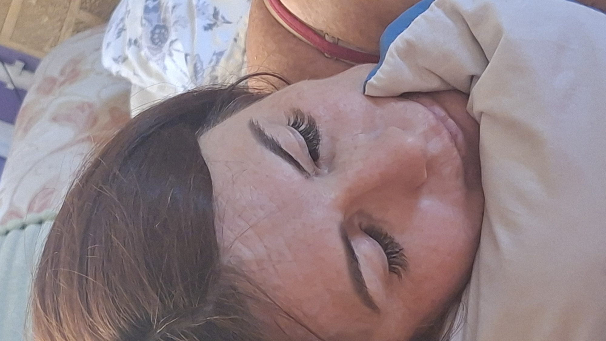 A woman with brown hair and long eyelashes is sleeping peacefully on a pillow. She is wearing a pink bra. The background is a bed with a floral patterned duvet. The scene appears to be indoors, possibly in a bedroom in Voinova, Moldova.
