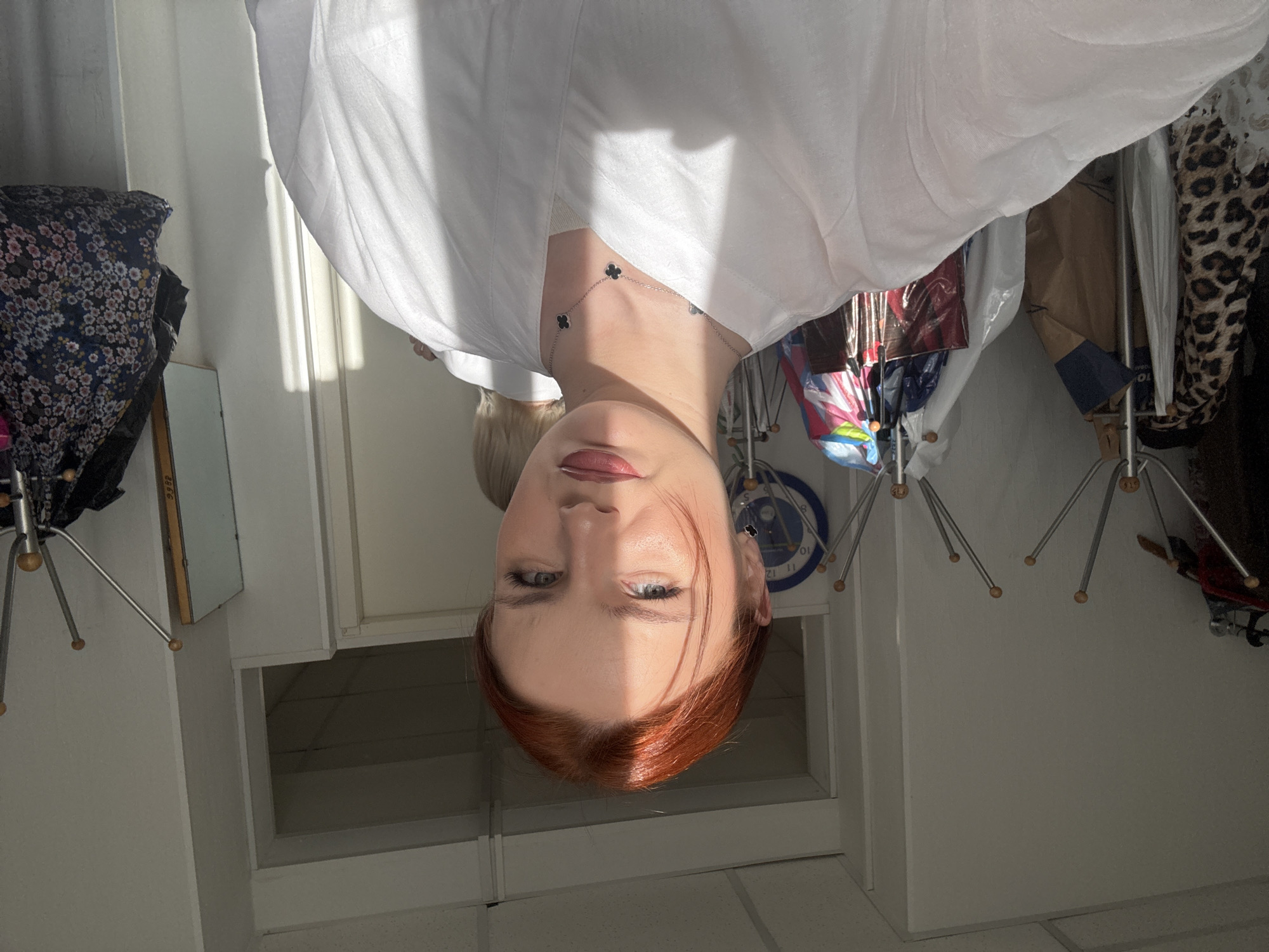 The image depicts a woman with red hair and fair skin, wearing a white shirt and a necklace with clover-shaped pendants. The background appears to be an indoor space, possibly a storage room or closet, with items such as clothing, bags, and what appears to be a drying rack. A blue and white clock is visible on the wall. Given the presence of typical Russian apartment elements like clothing drying racks and the architectural style, it is probable this is a building inside of Saint Petersburg, Russia. The photograph is taken from a low angle, making the subject appear to be looking down at the viewer.
