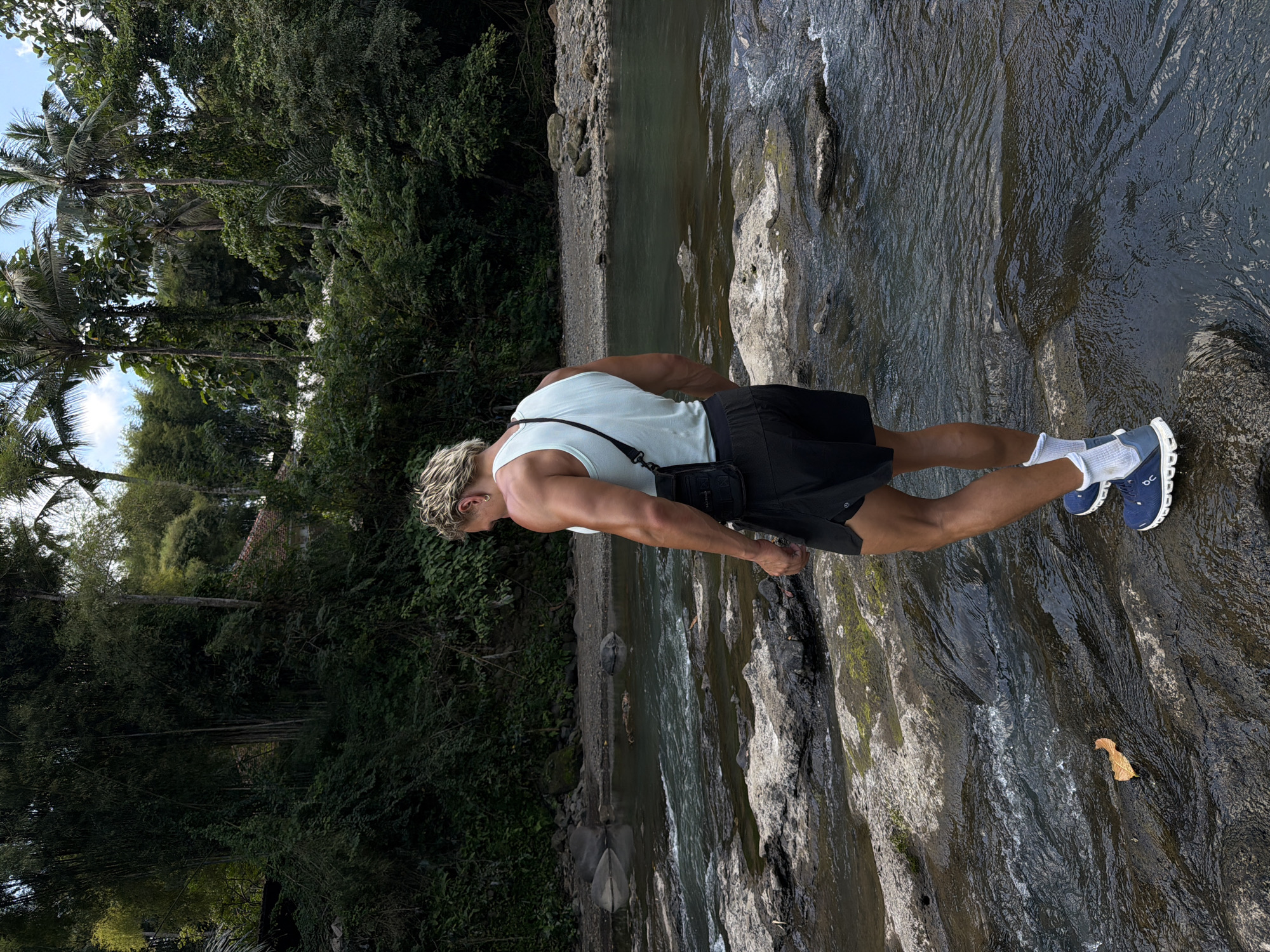 The image captures an individual standing on rocks in a riverbed, seemingly within a natural environment, possibly in Kerambitan, Indonesia. The person, likely a man with blond hair, is wearing a light-colored tank top, dark shorts, white socks, and blue sneakers. A black crossbody bag is also visible. The river flows past rocks, and dense foliage, including palm trees, lines the riverbank. The scene is bathed in daylight.