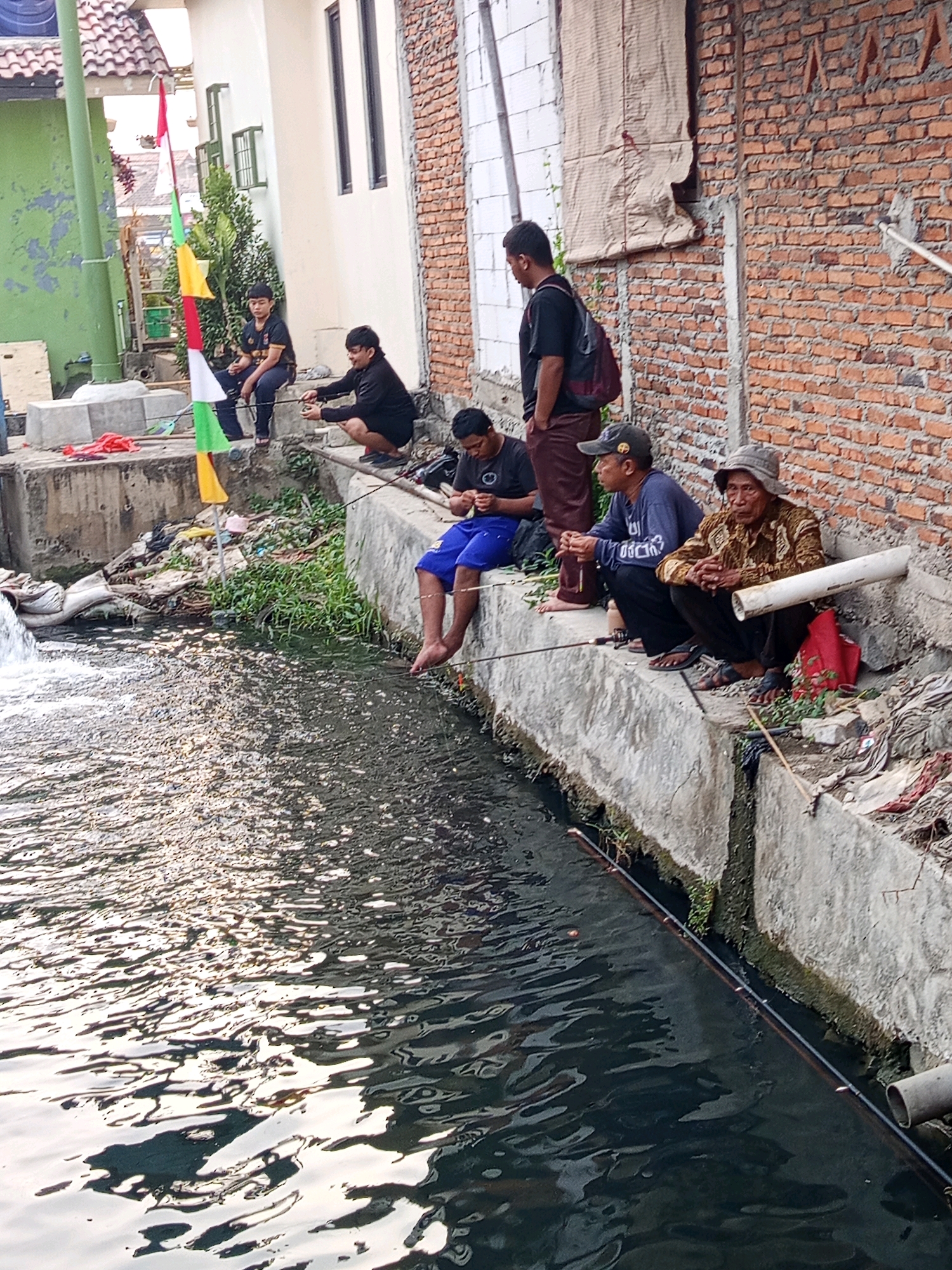 Six individuals are depicted fishing in a canal in the city of Karawang, Indonesia. The canal is lined with concrete walls, and the surrounding environment includes a brick building, a residential structure with a tiled roof, and vegetation. The water in the canal is dark and appears somewhat polluted, with visible debris along the banks.
From left to right, the first individual is a young boy sitting on the concrete embankment, holding a fishing rod. Next to him, a younger boy is crouching and also fishing. Further to the right, a man in a black shirt and blue shorts is sitting with his legs in the water, fishing. Beside him, another man is kneeling and holding a fishing rod. To the far right, an elderly man wearing a patterned shirt and a hat is seated and fishing, with a red bag at his side. Another man is standing in the background, facing away from the camera, wearing a black shirt and a backpack.
The scene suggests a casual outdoor activity taking place in a semi-urban environment. The presence of a waterfall or rapid in the canal on the left side of the image adds a dynamic element to the watery landscape. Flags, possibly decorative or celebratory, are also visible, attached to a pole near the left side of the frame.