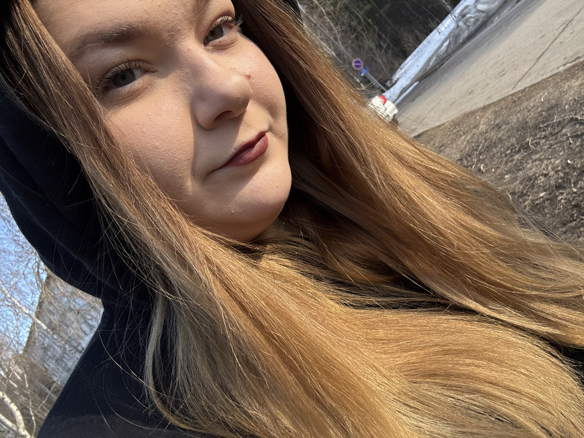 A close-up selfie shows a light-skinned woman with long, blonde hair, wearing a dark hooded garment. She is looking towards the left. The background reveals a street scene with a guardrail and foliage suggesting an outdoor location. Based on road signs and the general appearance of the environment, the location appears to be in the city of Chaikovsky, Russia.