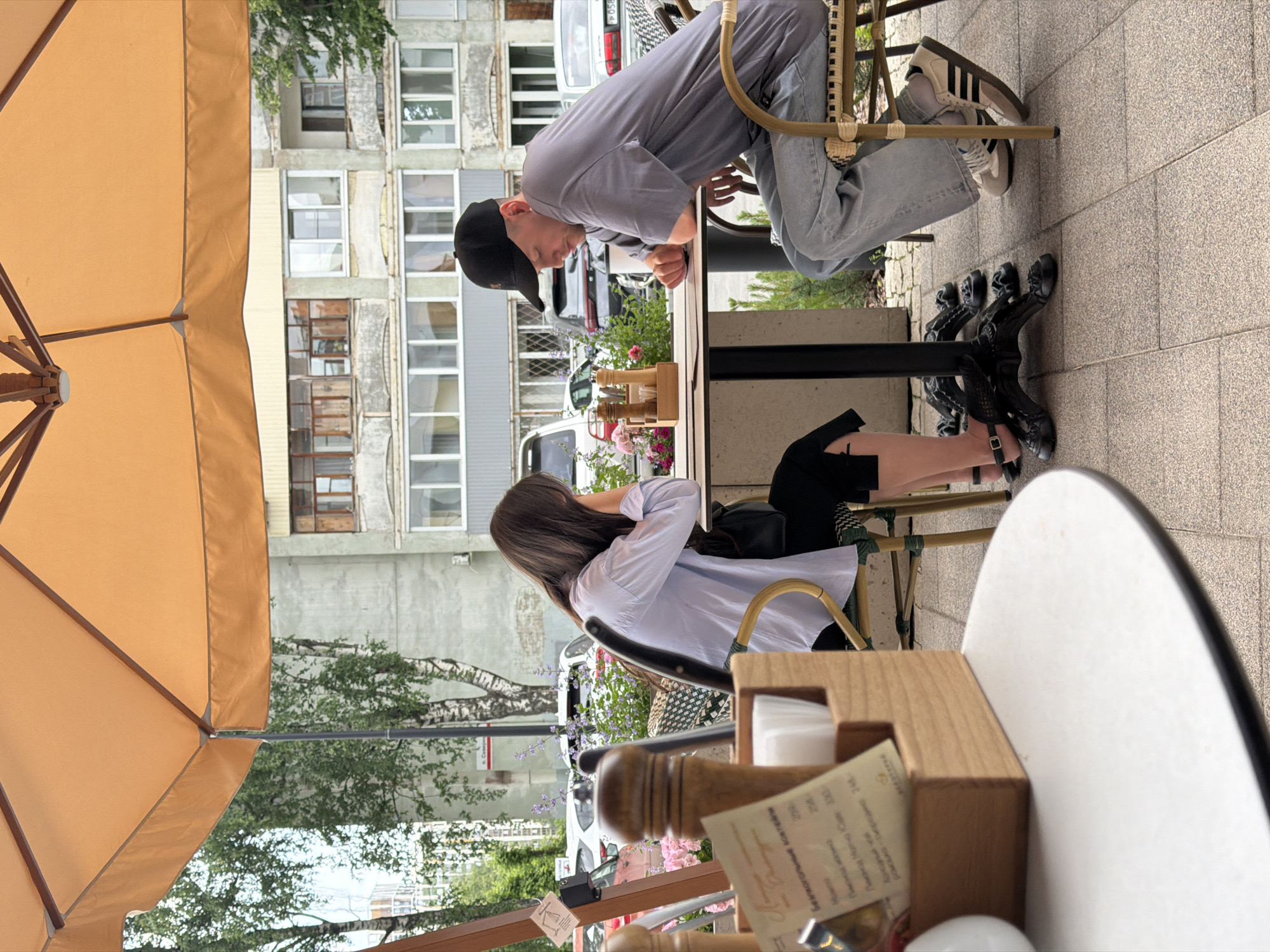 The image shows a man and a woman sitting at a table outside a cafe on a street in Izhevsk, Russia. The woman is wearing a light blue shirt and black pants, and the man is wearing a grey t-shirt and blue jeans, and a black baseball cap. They are both looking down at the table. There is a menu in a wooden holder on the table, as well as a salt and pepper shaker set. In the background, there are buildings, trees, and cars. The cafe appears to be located on a sidewalk next to a street. The sky is overcast.