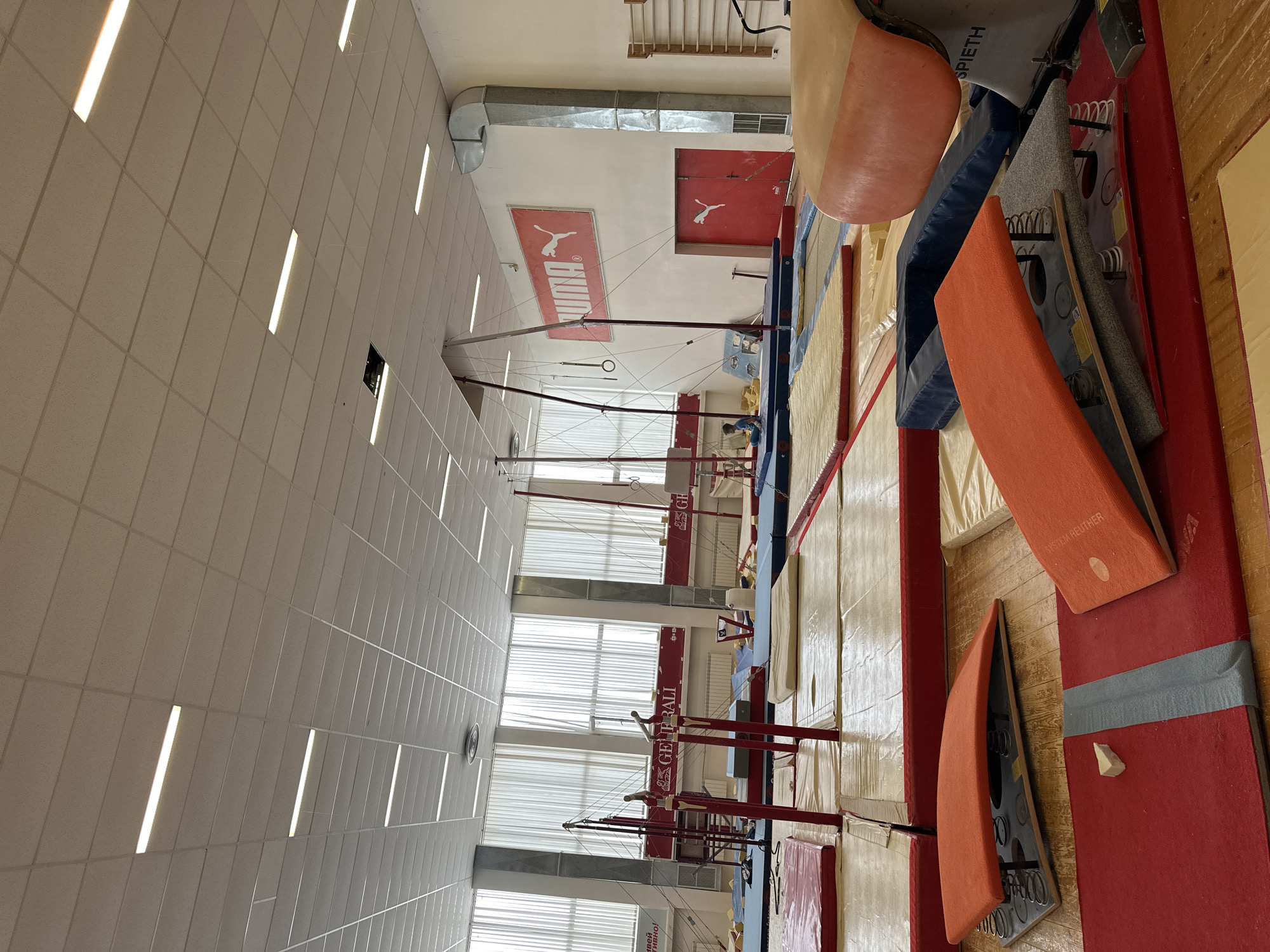 The image depicts the interior of a gymnasium in Sofia, Bulgaria. The gym features gymnastics equipment, including uneven bars, parallel bars, and mats. A "Puma" logo is visible on a wall. A row of windows provides natural light. The lighting system consists of fluorescent tube fixtures in a suspended ceiling. Gymnastics landing mats of various colors and sizes are stored against the walls. A gymnastics spring board is on the ground near the mats.