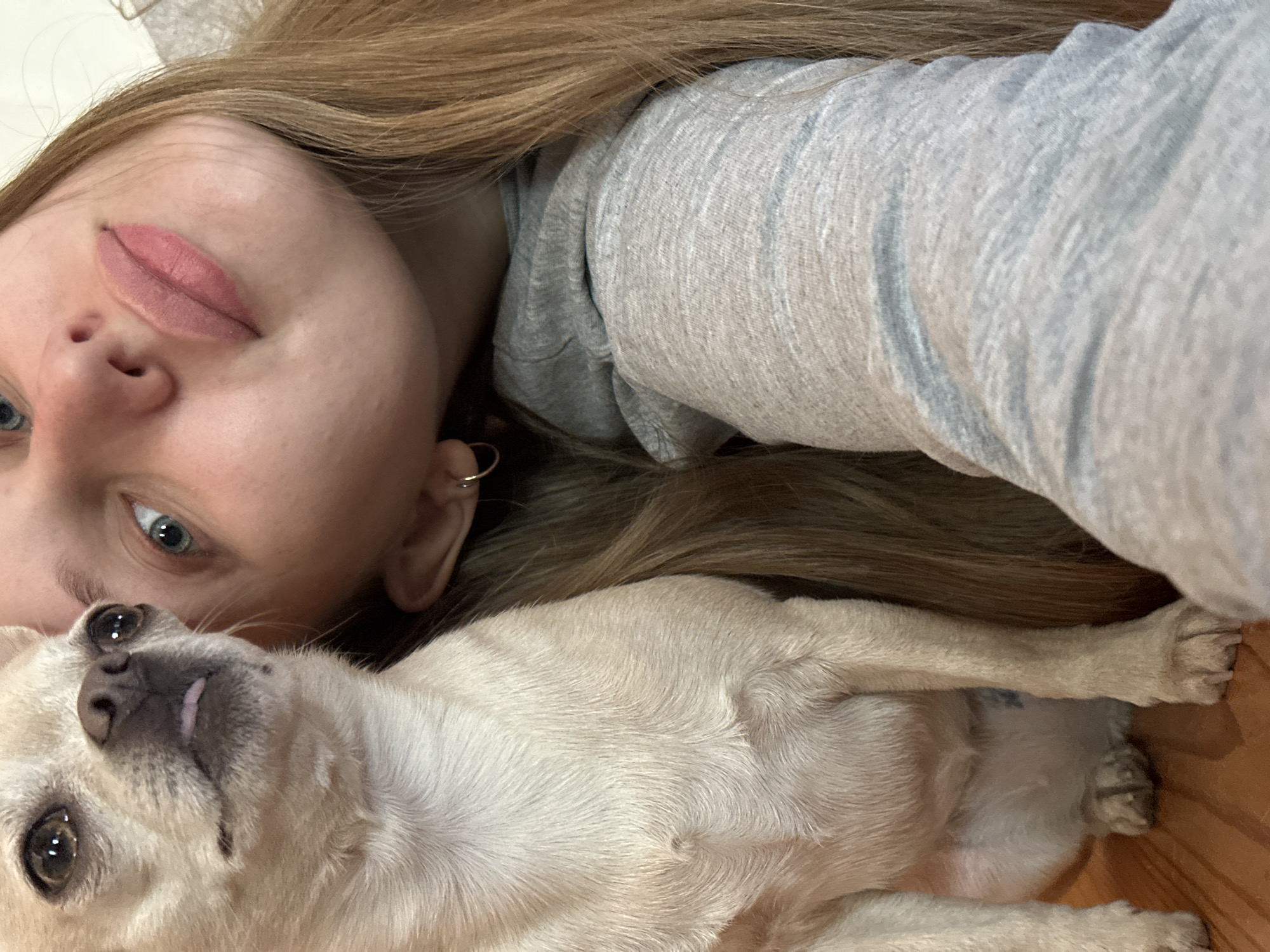 The image shows a woman and a dog lying down indoors. The woman is fair-skinned with blue eyes and light brown hair, wearing a gray sweatshirt. She is positioned with her head close to the camera, her face slightly tilted. The dog, a small, light-colored breed, rests beside her, appearing relaxed. The background is indistinct, but includes a wooden floor. There is no indication of this image being taken in the city of Chaikovsky, Russia.