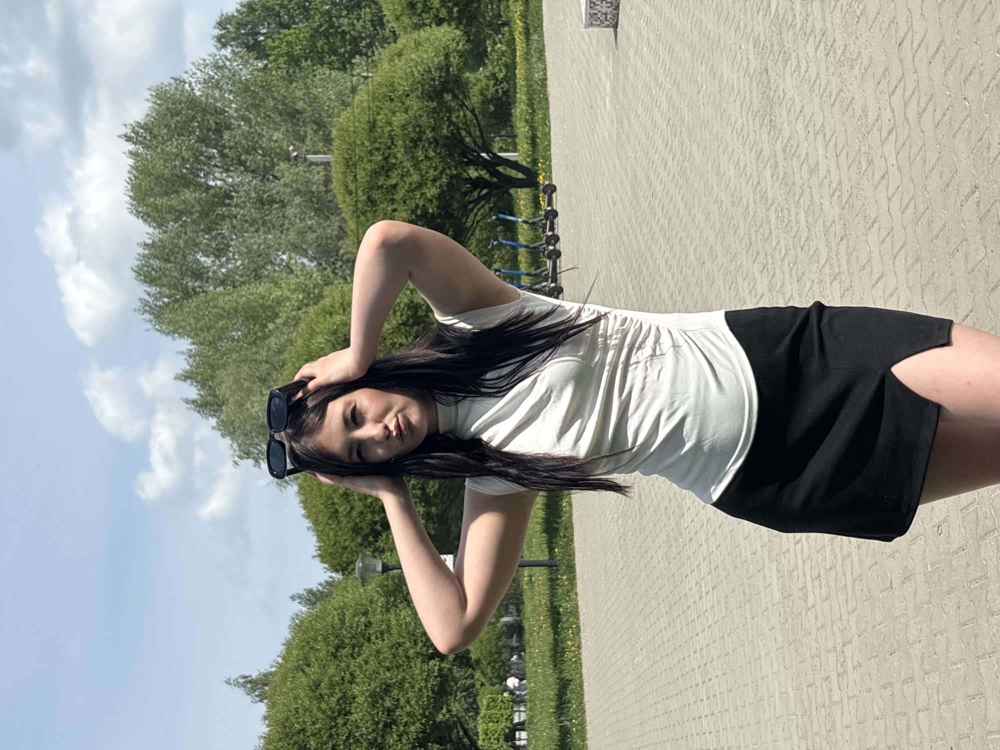 A young woman stands on a paved pathway outdoors. She is wearing a white t-shirt and a black skirt. Her dark hair falls over her shoulders, and she is wearing sunglasses on top of her head. Her hands are raised near her head. The pathway is made of interlocking concrete pavers. In the background are green trees and blue sky. The location is Chaikovsky, Russia.