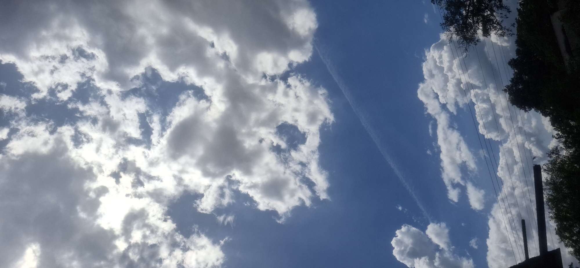 The image shows a sky with a mix of cumulus and stratocumulus clouds. A contrail stretches diagonally across the blue expanse between the clouds. The right side of the frame includes trees and what appears to be an old, disused structure with vertical elements, possibly chimneys or posts, and a few electrical wires. The location is inferred to be Vallecito, Venezuela. No people are visible.