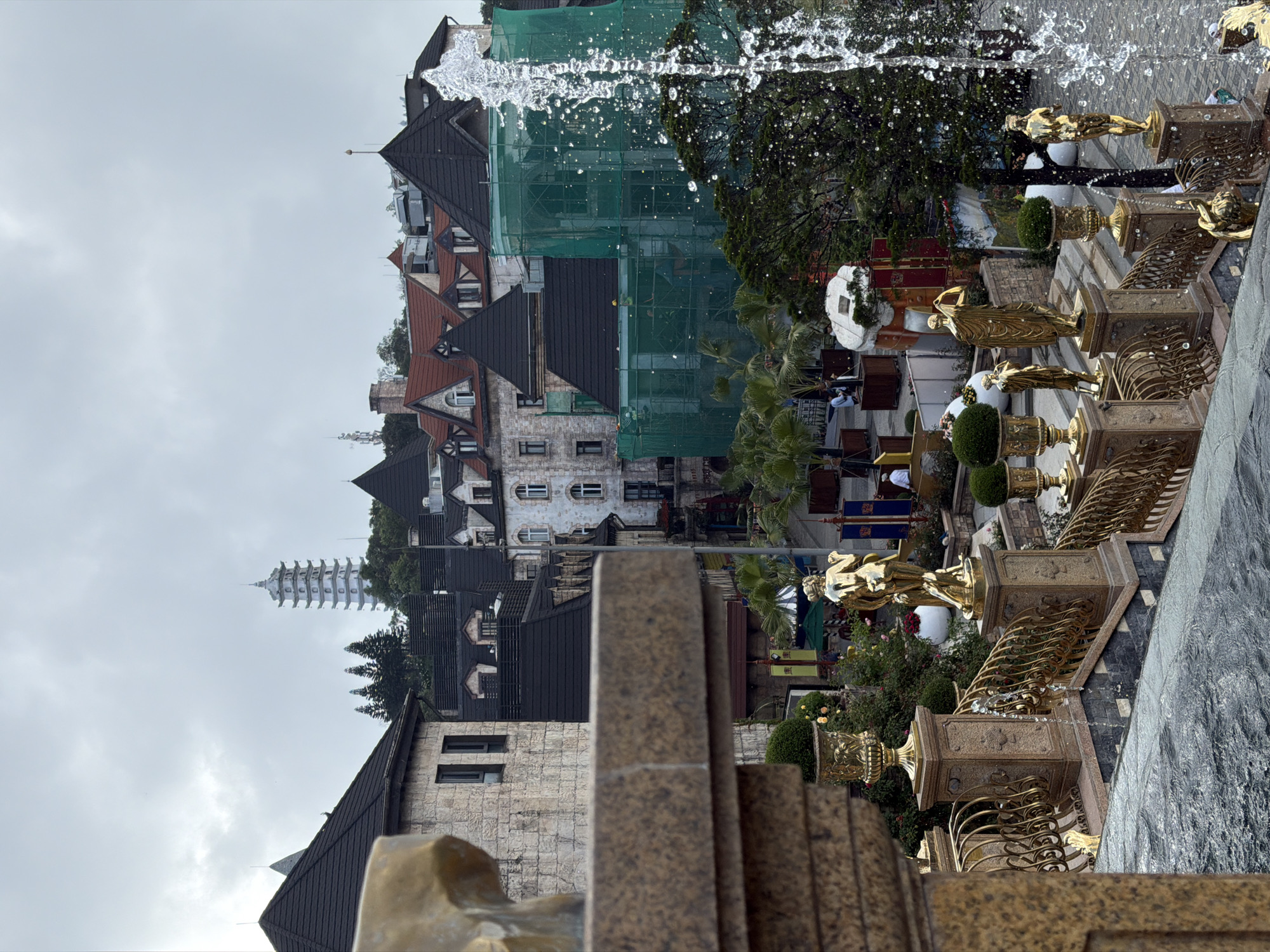 The image depicts a cityscape, possibly in Đà Nẵng, Vietnam, viewed from an elevated position with a stone and gold-colored fountain in the foreground. The fountain features golden statues and decorative elements. Beyond the fountain, there's a tiered arrangement of buildings with European-style architecture, including steeply pitched roofs and various architectural details. Scaffolding and green mesh cover parts of one building, suggesting ongoing construction or renovation. A tall, white, pagoda-like structure rises above the buildings. The sky is overcast. People are seen near the fountain and in the background among the buildings.