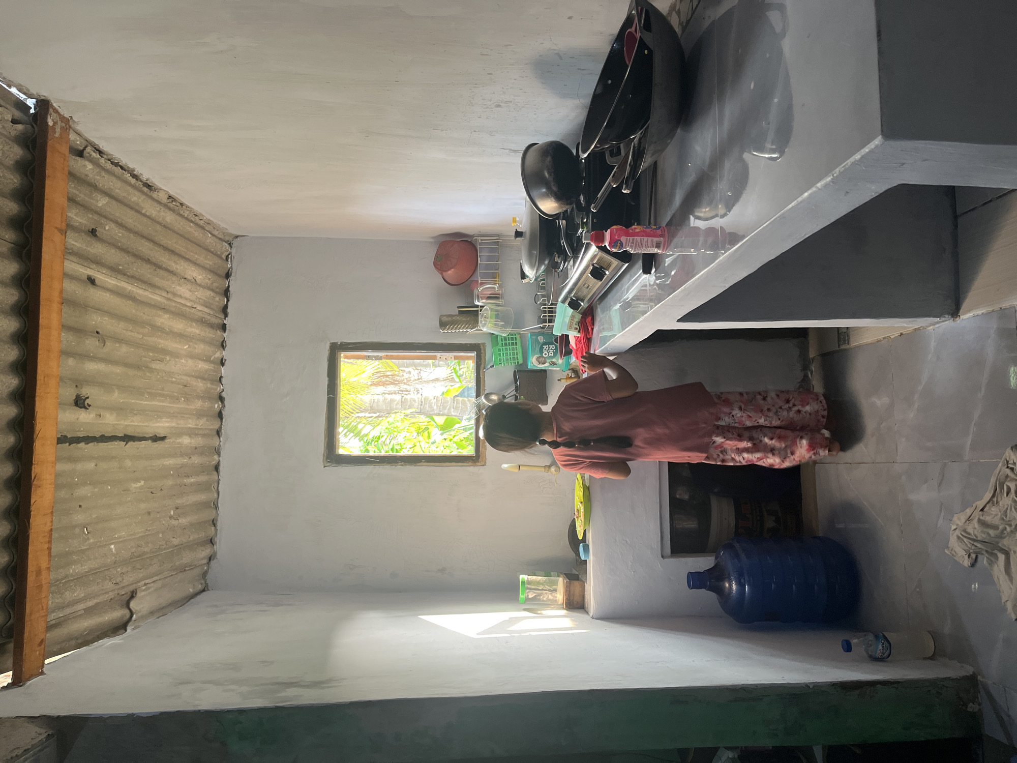 A young girl with dark hair in a braid, wearing a coral-colored t-shirt and floral-patterned pants, stands in a dimly lit kitchen in Laban, Indonesia. She is facing away from the camera, with her back to a small, open window that overlooks lush greenery and palm trees. The girl appears to be washing dishes at a concrete sink, with a green plate visible on the counter. The kitchen features a simple, unfinished aesthetic. The walls are painted a light gray, and the corrugated metal ceiling shows signs of wear. To the right of the girl, a grey concrete countertop holds cooking supplies, including pots, pans, and a gas stove. A large blue water dispenser is situated on the floor to her left, next to a partially visible white plastic bottle. The floor is tiled, and a piece of light-colored fabric lies on it in the foreground. The overall impression is of a humble, functional kitchen space.