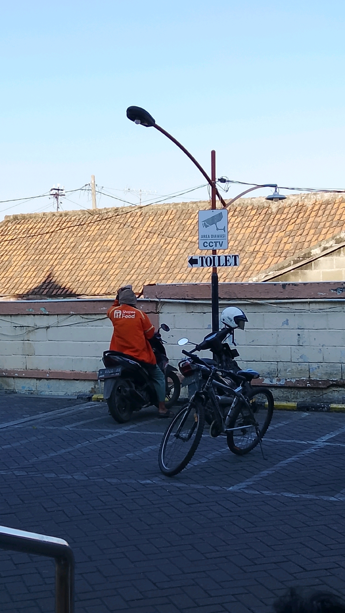 An outdoor scene in the city of Ngipik, Indonesia, features two individuals, one on a motorcycle and another on a bicycle, with a shophouse in the background. The shophouse has a terracotta tiled roof and a light-colored brick wall. A black street lamp with a single light bulb arcs over the scene. A traffic sign with an arrow points towards a "TOILET," with an additional inscription indicating "AREA DIAWASI CCTV."
The person on the motorcycle wears an orange jacket with a "Shopee Food" logo and a head covering, and is seated on a black motorcycle with a license plate that appears to read "W 6920 AM." The person on the bicycle wears a white helmet and a dark jacket and is seated on a black bicycle, positioned in front of the motorcycle. The ground is paved with dark interlocking bricks, with white painted parking lines.