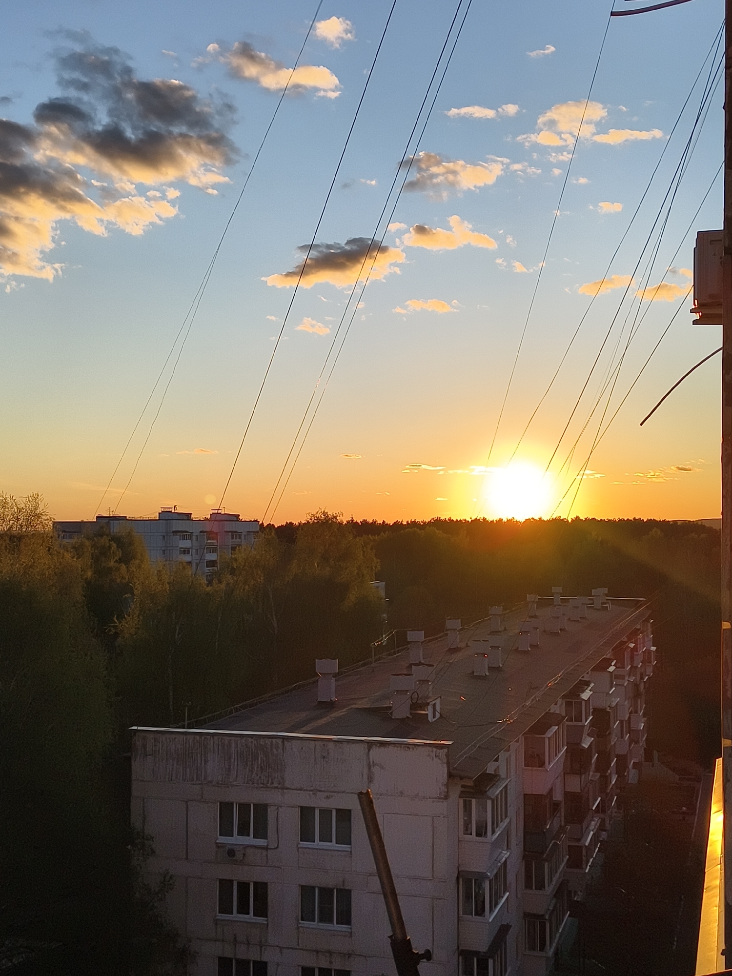 The image captures an elevated view of a cityscape at sunset. The sky is predominantly blue, streaked with orange hues near the horizon, where the sun is setting. Wispy clouds, tinted with gold, are scattered across the sky. Power lines are visible, crisscrossing the upper portion of the frame.
Below the horizon, a dense line of trees forms a silhouette against the bright sunlight. In the foreground, several buildings are visible. One building is pale, likely concrete, with multiple windows and a flat roof dotted with vents. Balconies protrude from its sides. Another taller building can be seen further in the distance, behind the trees.
The lighting indicates a late afternoon or early evening scene. The buildings and trees are partially obscured by shadows. The overall mood is peaceful and serene. The location is a city, but it is not identifiable from the image.