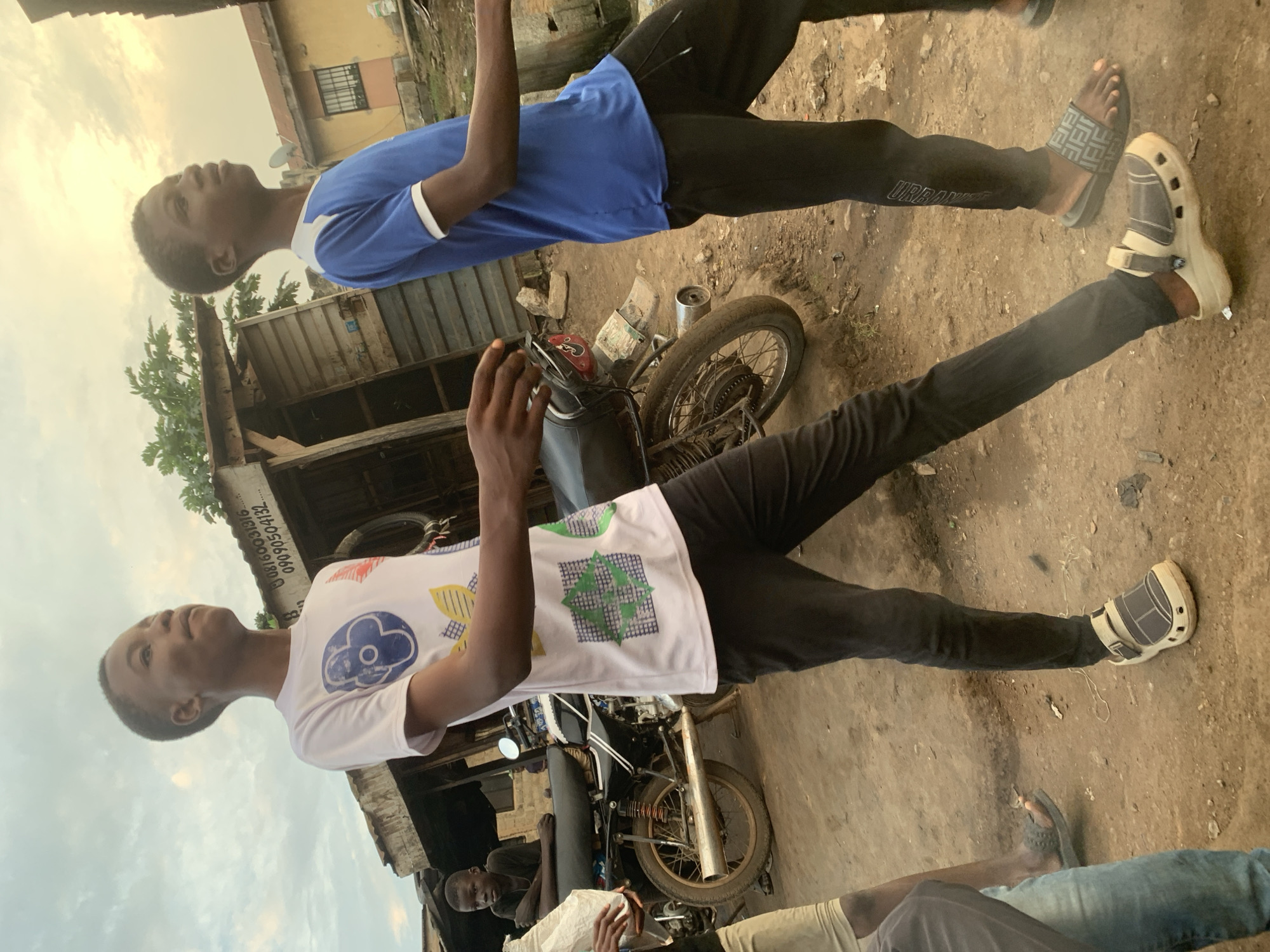 In the city of Shagamu, Nigeria, two young boys walk on a dirt road. The boy on the left wears a white t-shirt with a colorful graphic print and black pants, with white and grey clogs on his feet. The boy on the right wears a blue t-shirt with white trim and black pants, with black and grey slides on his feet. Behind them, a motorcycle is parked next to a makeshift stall constructed of corrugated metal and wood. The stall has signage that includes a phone number. The ground is dusty and uneven. The sky is overcast with soft light.