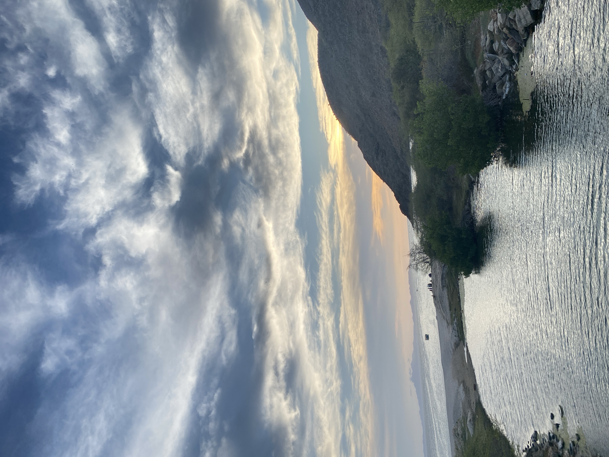 The image shows a scenic view of the coast near Juan Griego, Venezuela, capturing a blend of natural elements and human presence.
The sky is expansive, with a mix of blue and grey clouds, suggesting either early morning or late afternoon light. The sun's golden light peeks out from behind a large landmass, possibly a mountain or large hill, that dominates the midground of the image. The coastline is lined with vegetation, including trees and bushes. A calm body of water reflects the sky.
Small figures of people are visible on the beach. Some are standing and some appear to be walking. Further offshore, at least one boat is visible. The image is likely taken from an elevated position, providing a panoramic perspective of the scene.