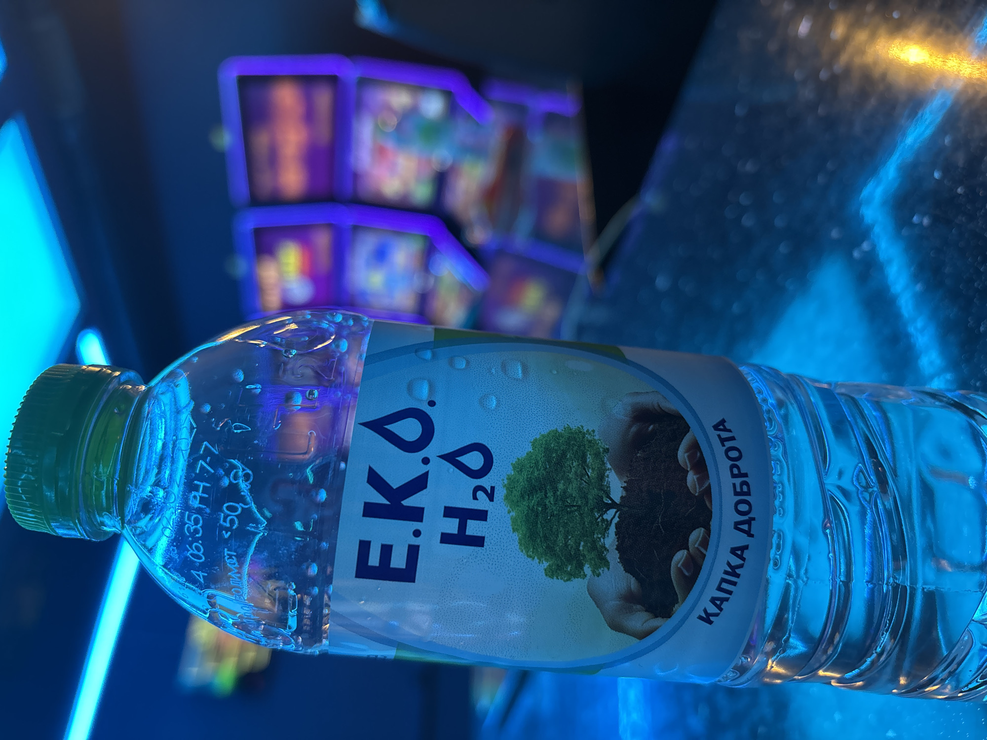 A horizontal, medium shot depicts a bottle of E.K.O. H2O water. The water bottle is positioned in front of a blurry slot machine. The bottle's label displays the words "E.K.O. H2O," along with an image of a tree growing in soil held within two hands. Text on the label reads "КАПКА ДОБРОТА," which translates to "DROP OF GOODNESS." The location is potentially in the city of Pleven, Bulgaria, as E.K.O. H2O is a brand produced in Bulgaria. The scene is illuminated with blue light. The water bottle is slightly angled, with the cap on the left.