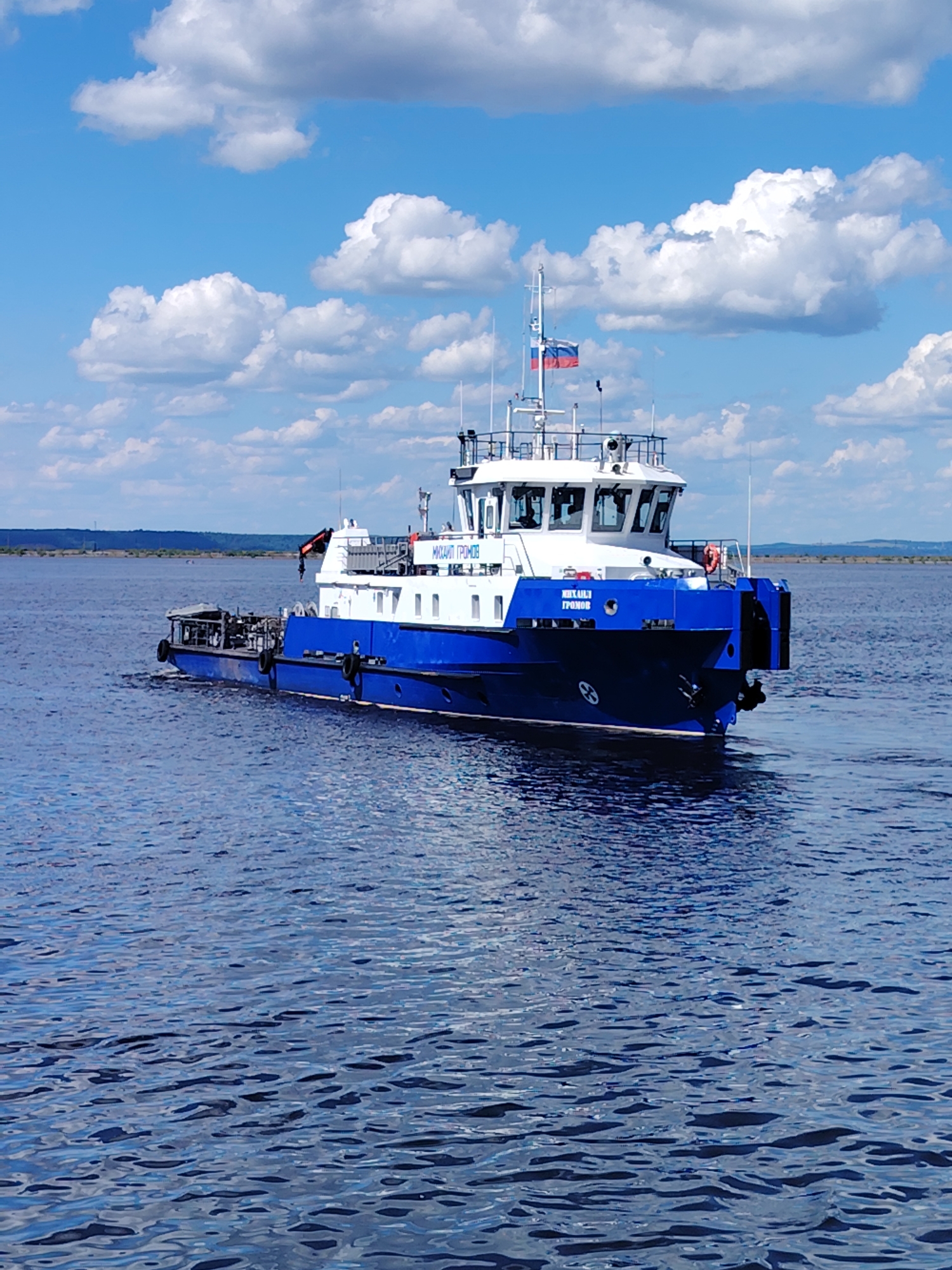A blue and white vessel, named "МИХАИЛ ГРОМОВ", is navigating on a body of water. The vessel has a white superstructure with a bridge at the front, and a blue hull. A Russian flag is visible on a mast on the superstructure. The vessel is a tugboat or a service boat, possibly in a port or on a river. The water is blue with some ripples, indicating a sunny day with clouds in the sky. The background shows a distant, hazy shoreline with trees and some structures. The image appears to be taken from another boat or from the shore. The location is the Kama River in Chaikovsky, Russia, given the presence of the "MIHAIL GROMOV" vessel, which is a known vessel operating in that region.