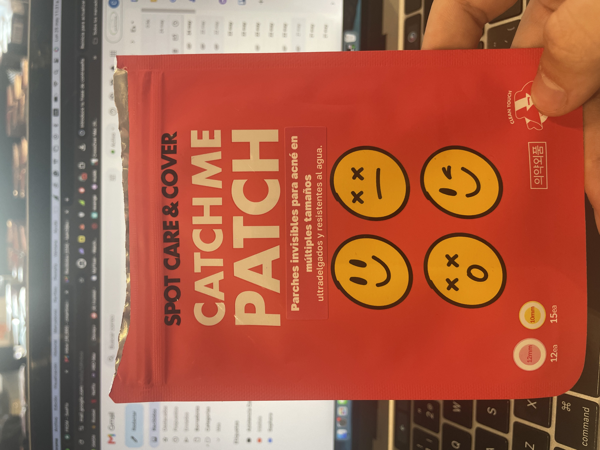 The image shows a red package of "Spot Care & Cover Catch Me Patch," acne patches in multiple sizes, held by a person's right hand. The package features four yellow smiley face designs with different expressions. Text on the package includes "Parches invisibles para acné en múltiples tamaños ultradelgados y resistentes al agua," indicating they are invisible acne patches in multiple sizes, ultra-thin, and water-resistant. Additional text includes size indications of 12mm (12ea) and 10mm (15ea). The package also includes a "Clean Touch" symbol, along with Korean text "의약외품."
The package is positioned over a laptop keyboard. In the background, a blurred laptop screen displays a Gmail inbox. This scene takes place indoors. There is no indication this image was taken in Mexico City, Mexico.