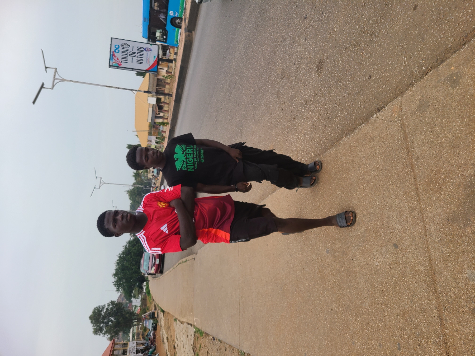 This image, rotated ninety degrees clockwise, captures two young men standing on a paved sidewalk adjacent to a road in what appears to be an urban or semi-urban area in Jos, Nigeria, under an overcast sky during daytime.
The young man on the left is wearing a red football jersey, likely a Manchester United kit with the Adidas logo, black shorts, and black sandals. His arms are crossed, and he is looking slightly to the right of the frame. The young man on the right is dressed in a black t-shirt that prominently displays a green eagle graphic and the text "NIGERIA SOCCER CAMP 2023 1 CORINTHIANS 9:26 BATCHES". He wears black trousers and black sandals, also with his arms crossed, and is looking towards the camera. Both individuals appear calm and posed for the photograph.
In the background, a road is visible with a blue and white bus partially cropped on the right. A tall street light pole stands out against the pale sky. Most notably, a large political billboard is situated on the far side of the road, displaying the slogan "TINUBU — OR — NOTHING" with a stylized symbol and the words "LAGOS STATE CHAPTER". An image of a man's face, presumably Bola Ahmed Tinubu, is also on the billboard. Various low-rise buildings, smaller vehicles, and some trees complete the street scene.