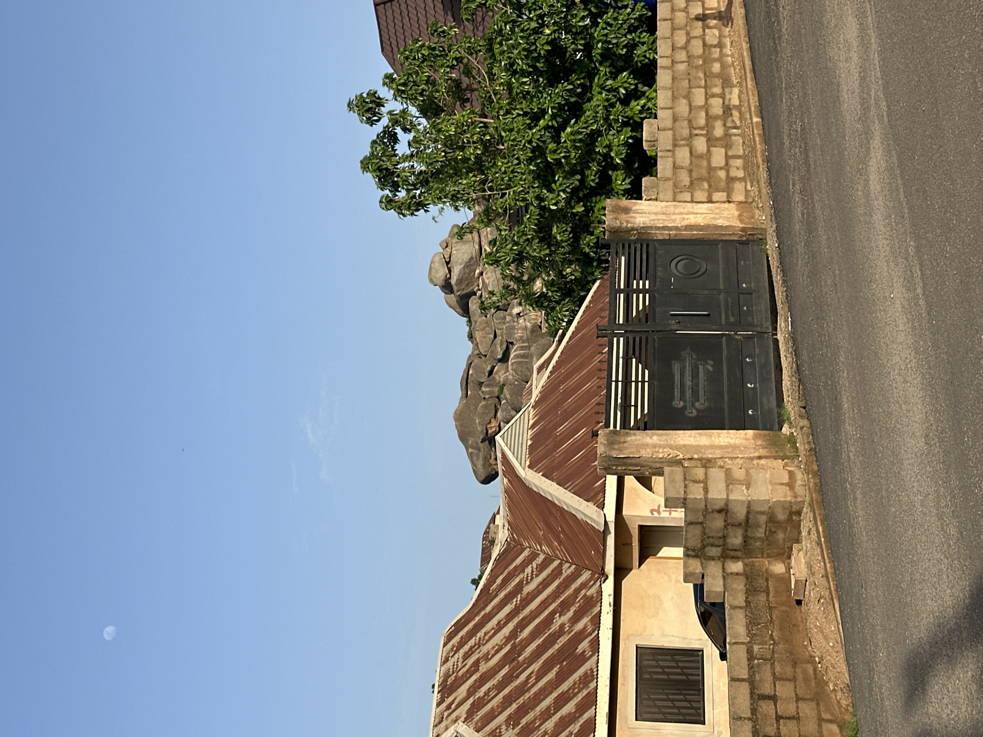 The image depicts a house with a corrugated metal roof in Jos, Nigeria. The house has a yellow stucco exterior and a dark metal gate. A paved road runs alongside the property, separated by a low brick wall. Lush green trees and large rock formations are visible behind the house, suggesting a natural, possibly hilly, landscape. The sky is clear and blue, with a faint crescent moon visible. The sunlight suggests it is daytime, likely late afternoon or early morning due to the angle of the shadows. There is no visible text in the image. The scene appears peaceful and residential, with no people or specific activities evident.