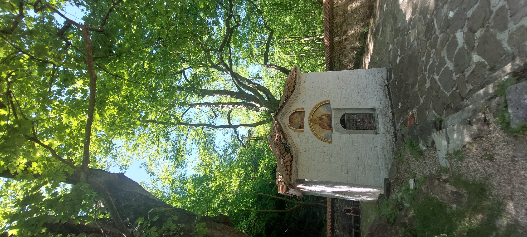 The image shows a small, white chapel with an orange tile roof in Бачково, Bulgaria. The chapel is adorned with religious paintings above a window and stands next to a stone wall. Trees surround the structure, creating a shaded, natural environment. A cross is visible on the roof of the chapel.