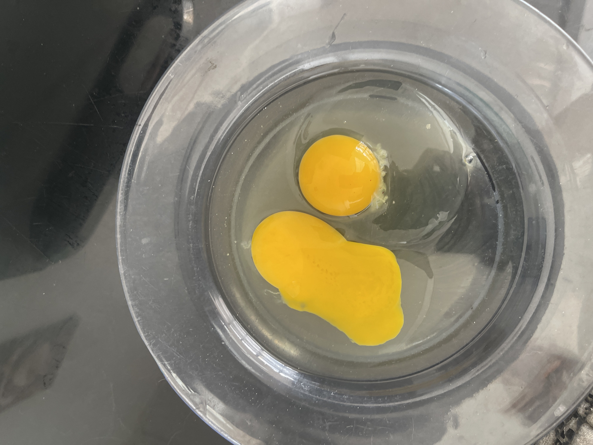 Two egg yolks are visible in a clear glass bowl on a dark, reflective surface. The yolk on the left is elongated and appears to be partially broken, with a small piece separated from the main body. The yolk on the right is round and intact. The background surface has some scuff marks and reflections. The image appears to be taken from a top-down perspective.
