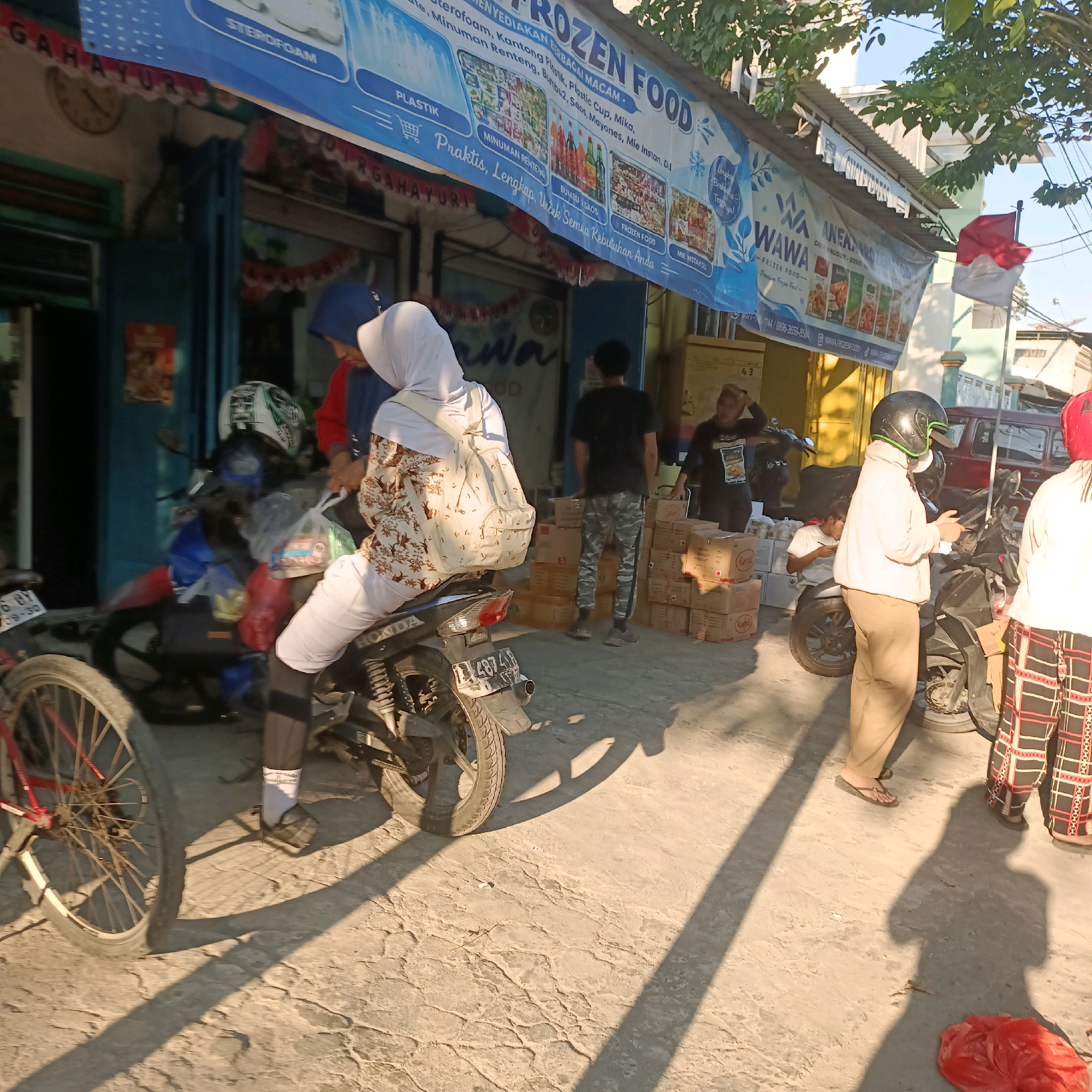 The image depicts a street scene in Surabaya, Indonesia, with a focus on a storefront displaying a banner for "FROZEN FOOD" and "WAWA FROZEN FOOD". The storefront appears to be a small shop or market. In the foreground, several people are visible, some of whom are near motorcycles. One person, wearing a hijab and a patterned top, is sitting on a motorcycle, holding a plastic bag. Another person in a hijab and white clothing is standing nearby. A person wearing a black shirt and camouflage pants is standing further back, near stacks of cardboard boxes. The environment is an outdoor setting with sunlight casting long shadows, suggesting it is daytime. The ground is paved with concrete. Other elements include a bicycle wheel, a red bag on the ground, and a red and white Indonesian flag flying on a pole in the background. The overall impression is a bustling street scene outside a retail establishment.