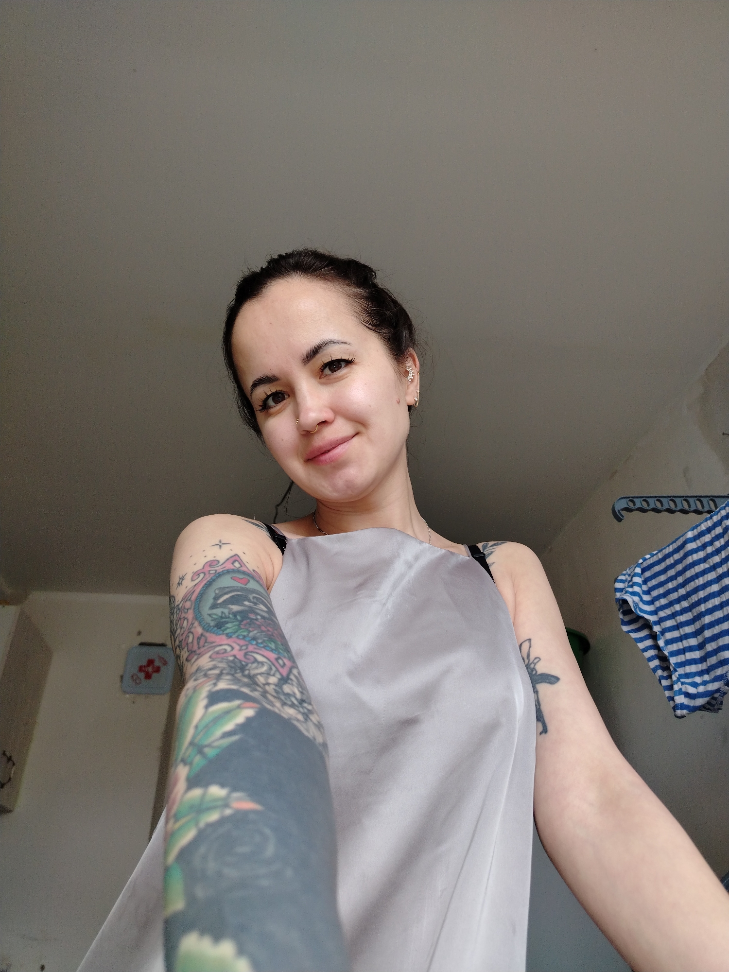 A young adult woman is captured in a close-up, upward-angled selfie, smiling faintly at the camera. She has dark hair pulled back, prominent eyebrows, and visible eyelashes. Her left nostril features a ring piercing, and her left ear has multiple stud and small hoop piercings. She wears a light grey, satin-like sleeveless top, with a dark bra strap visible on her right shoulder.
Her left arm, extended towards the camera and appearing prominently in the foreground, is covered with an elaborate, colorful full-sleeve tattoo featuring intricate designs that include floral motifs and what appear to be mythological or traditional Japanese elements. Her right upper arm also has a darker, simpler tattoo, partially visible beneath her top, which resembles a stylized object, possibly a rifle.
The setting appears to be an indoor room, likely a casual home environment in Noviy, Russia, judging by the background. The walls are a light, neutral color, appearing somewhat unfinished or unpainted. To the right, a blue plastic clothes hanger or rack holds a blue and white striped cloth. To the left, a small, light blue square object with a red floral or cross-like design is mounted on the wall. The lighting suggests it is daytime. No text is visible in the image.