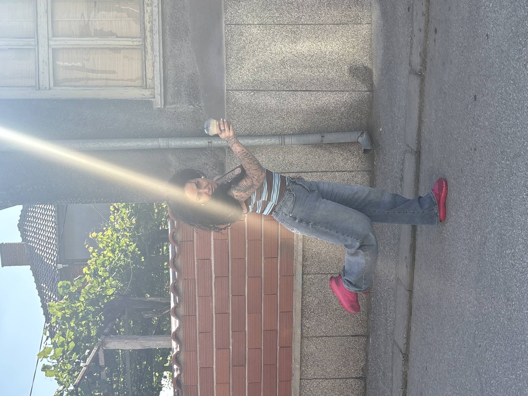 An outdoor, sunlit scene in the city of Rakovski, Bulgaria, depicts a female subject posing on an asphalt street. The subject, with dark, curly hair, wears a horizontally striped crop top (brown, light blue, and white), light blue loose-fitting jeans, and bright pink sneakers. She is standing on her left leg with her right leg bent at the knee and is holding a clear plastic cup containing a light-colored beverage in her extended right hand. Visible tattoos cover both of her arms. A prominent diagonal lens flare from the sun traverses the upper-left and central portions of the image.
The background on the left features a low red brick wall with a textured grey base, behind which dense green foliage, consistent with grapevines on a wooden trellis, is visible under a clear blue sky. A building with a tiled roof and a chimney is situated behind the foliage. To the right of the subject, a plain, light grey building wall displays a vertical grey drainpipe. An old, light-colored wooden window frame with obscured panes is visible on this grey wall in the upper right. The foreground consists of a dark grey asphalt road surface with a curb separating it from the adjacent walls.