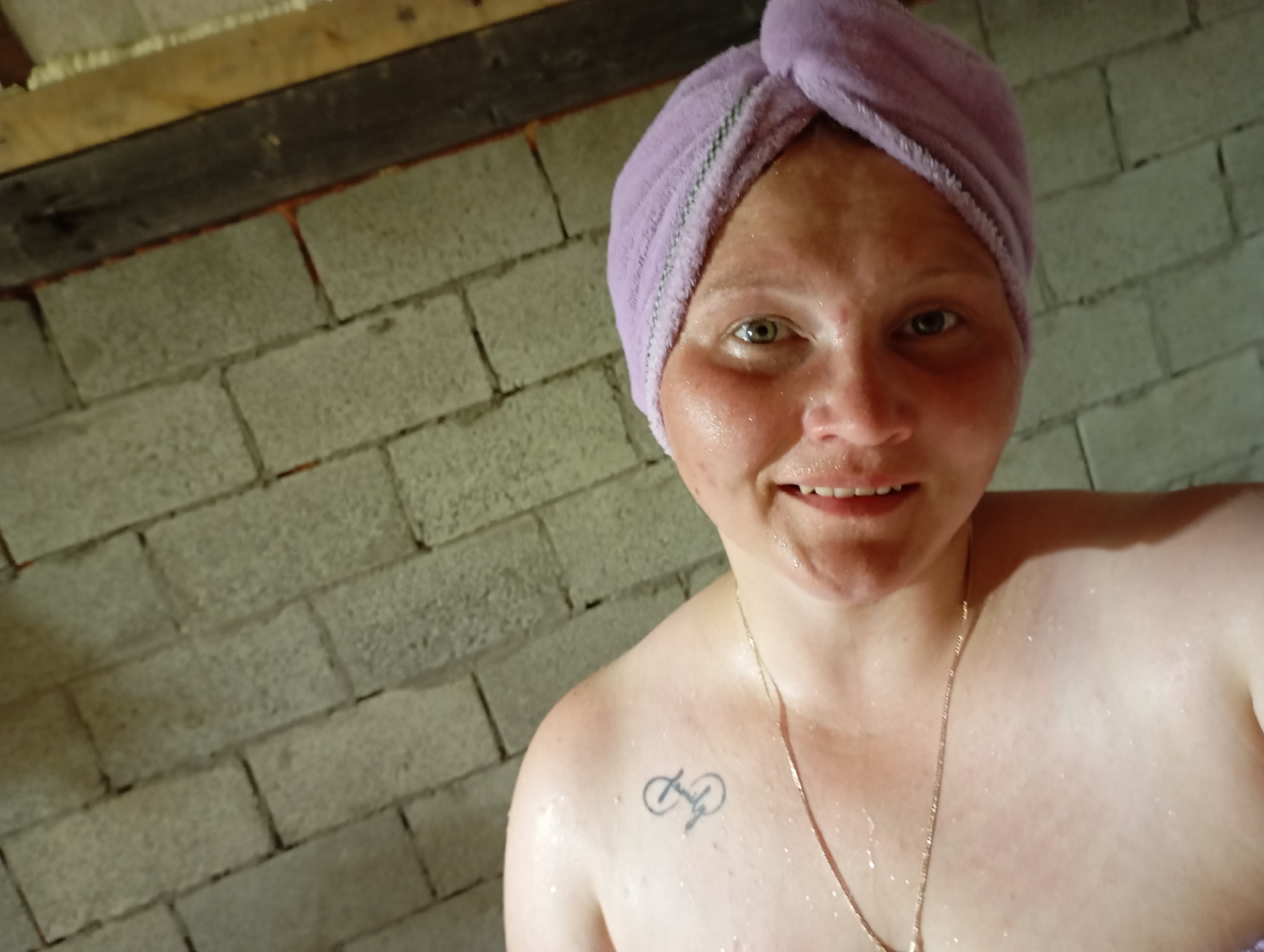 A person is shown from the chest up, wearing a purple towel on their head and a necklace. The person has a tattoo on their left shoulder. The background appears to be a brick wall, and there are wooden beams visible at the top of the image. The image is a close-up shot, suggesting it was taken by the person themselves. The lighting is dim, and the overall impression is that of a casual, personal photograph. The location of the image is not discernible from the visual cues present.