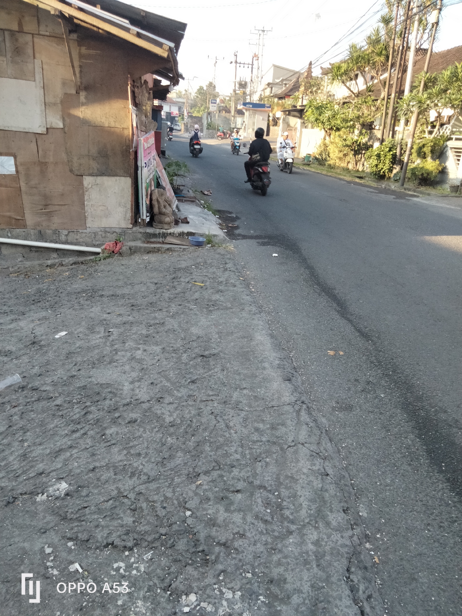 The image depicts a street scene in Denpasar, Indonesia, during daylight. The foreground is dominated by a cracked, gray, somewhat uneven ground surface, possibly a sidewalk or roadside area, with some scattered debris. A white pipe runs horizontally across this area.
The midground shows a paved asphalt road with several motorcycles and their riders. On the left side of the road, there is a small, weathered building with a corrugated metal roof. The building's walls appear to be constructed from various wood panels. Near the building, a sign is visible, along with a small statue, possibly of a deity.
In the background, the street continues, lined with houses and lush green vegetation, including palm trees. Overhead, electrical wires are strung between poles. The sky is clear and bright, suggesting sunny weather. A few people are seen riding motorcycles on the road, engaged in transit. One person in the midground is wearing a black shirt and pants, while others are in lighter clothing. The overall impression is of a typical urban street in a tropical climate.