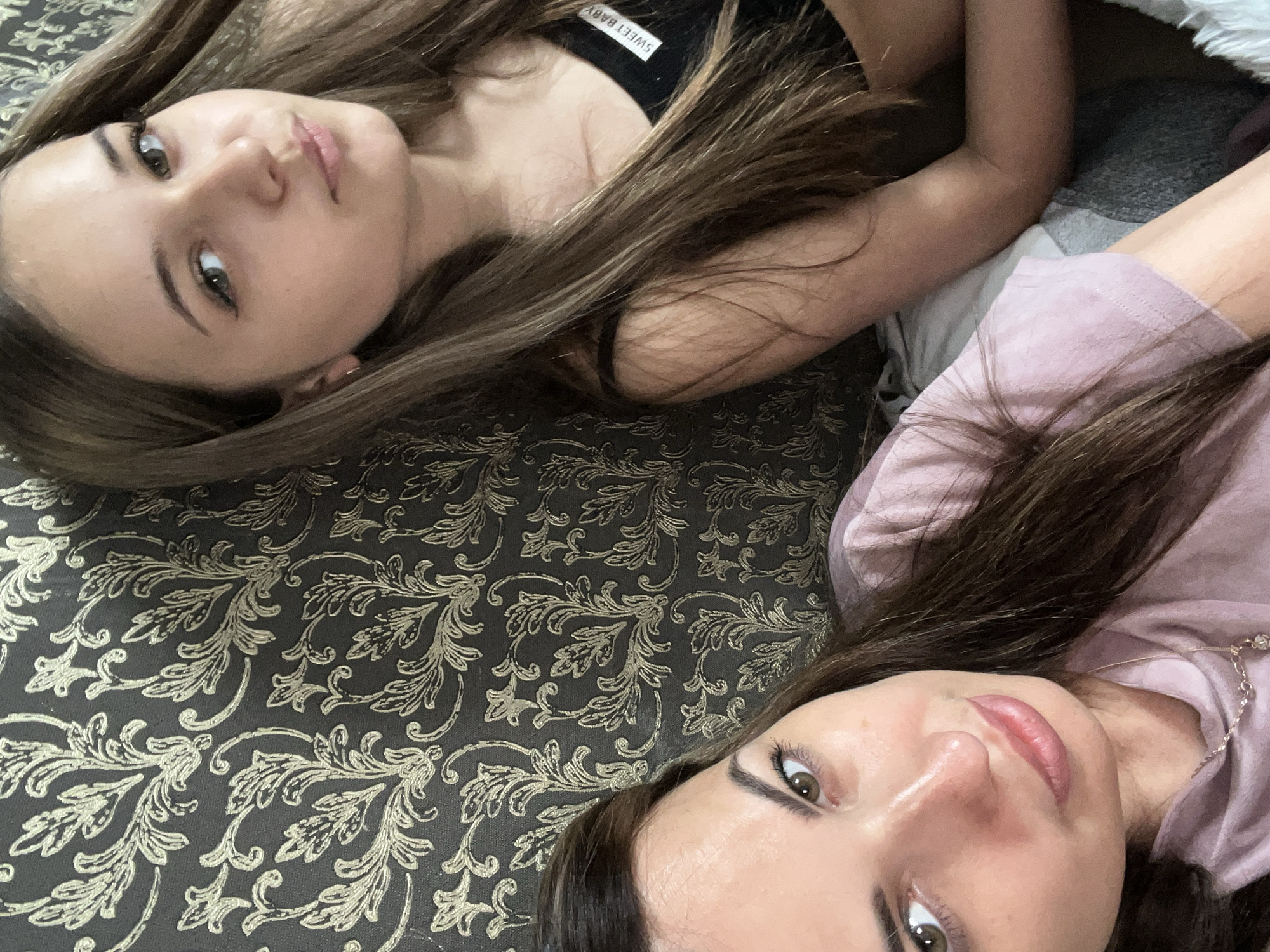 Two young women are pictured in a close-up shot, leaning against a patterned wallpapered surface. The woman on the left is in the foreground, looking directly at the camera with her head tilted slightly. She has brown hair and is wearing a pink shirt and a necklace. The woman on the right is partially visible, also looking towards the camera, with long brown hair and wearing a black top with the words "SWEET BABY" printed on it. The wallpaper behind them features an ornate, damask-like pattern in shades of dark gray and gold. The lighting is soft, suggesting an indoor setting. There is no discernible city or specific location information available from the image itself.