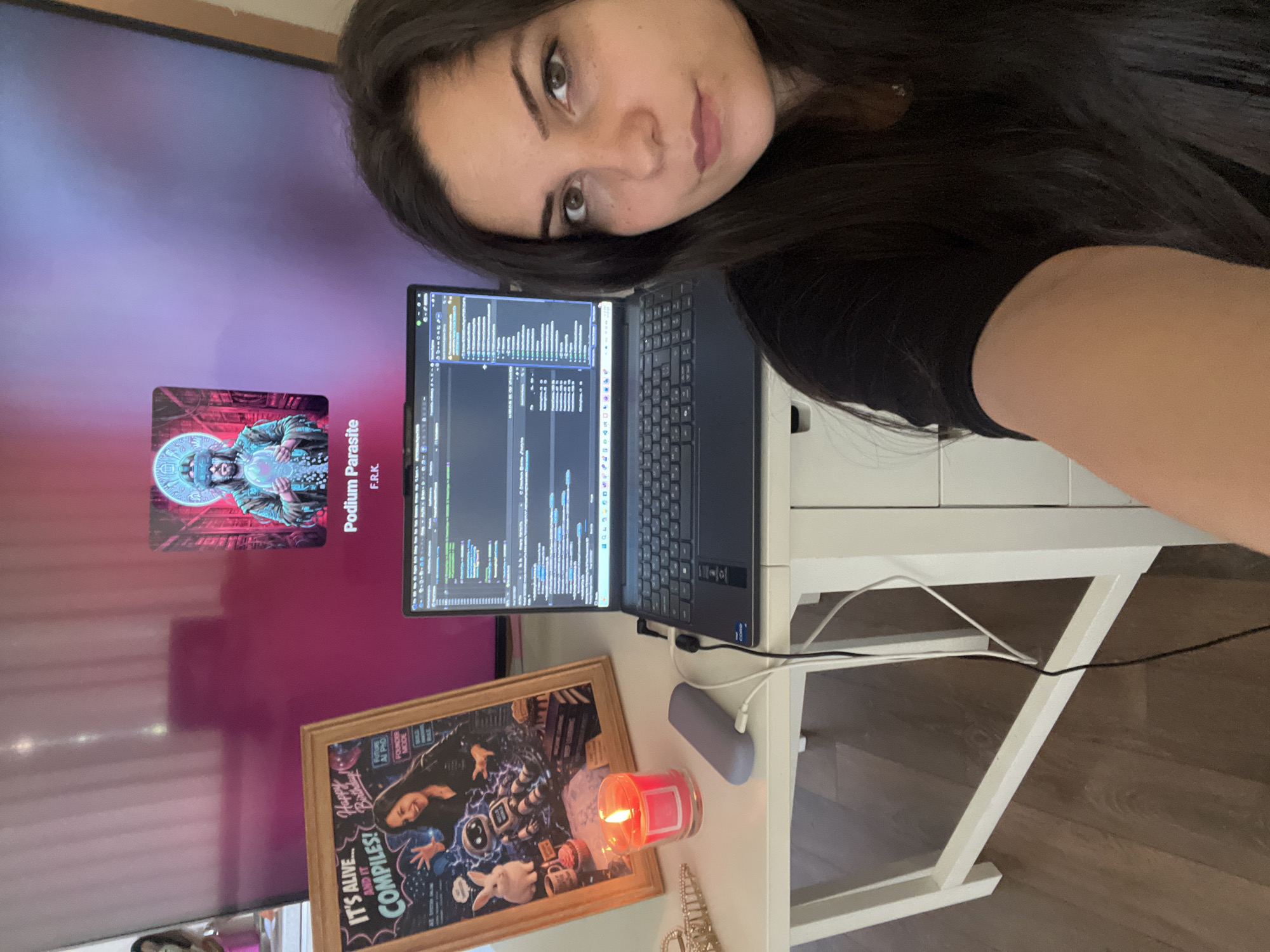 A woman with long dark hair is taking a selfie in a brightly lit room, with a laptop open on a white desk in front of her. The laptop screen shows lines of code. To the left of the laptop, a framed poster with the words "IT'S ALIVE... AND IT COMPILES! Happy Birthday!" is displayed, featuring an illustration of a woman and a robot. A lit candle in a red glass jar sits next to the poster, and a grey charging device is plugged into the laptop. In the background, a large television screen displays an image of a person wearing VR goggles, with the text "Podium Parasite F.R.K." below it. The overall impression is a home office or workspace. The location is not determinable from the image.